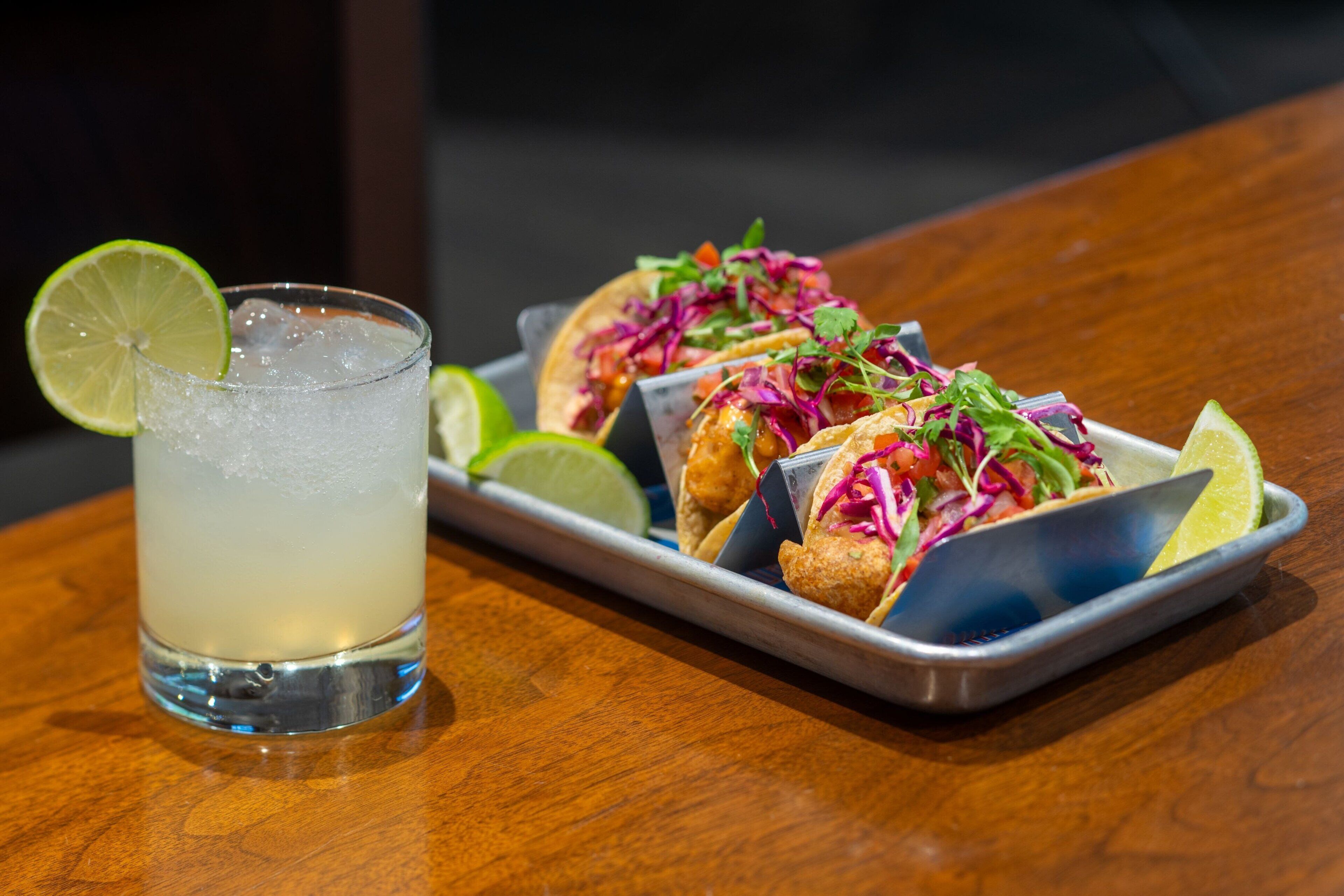 Una bandeja de tacos con una cuña de lima y un vaso de una bebida transparente sobre una mesa de madera.