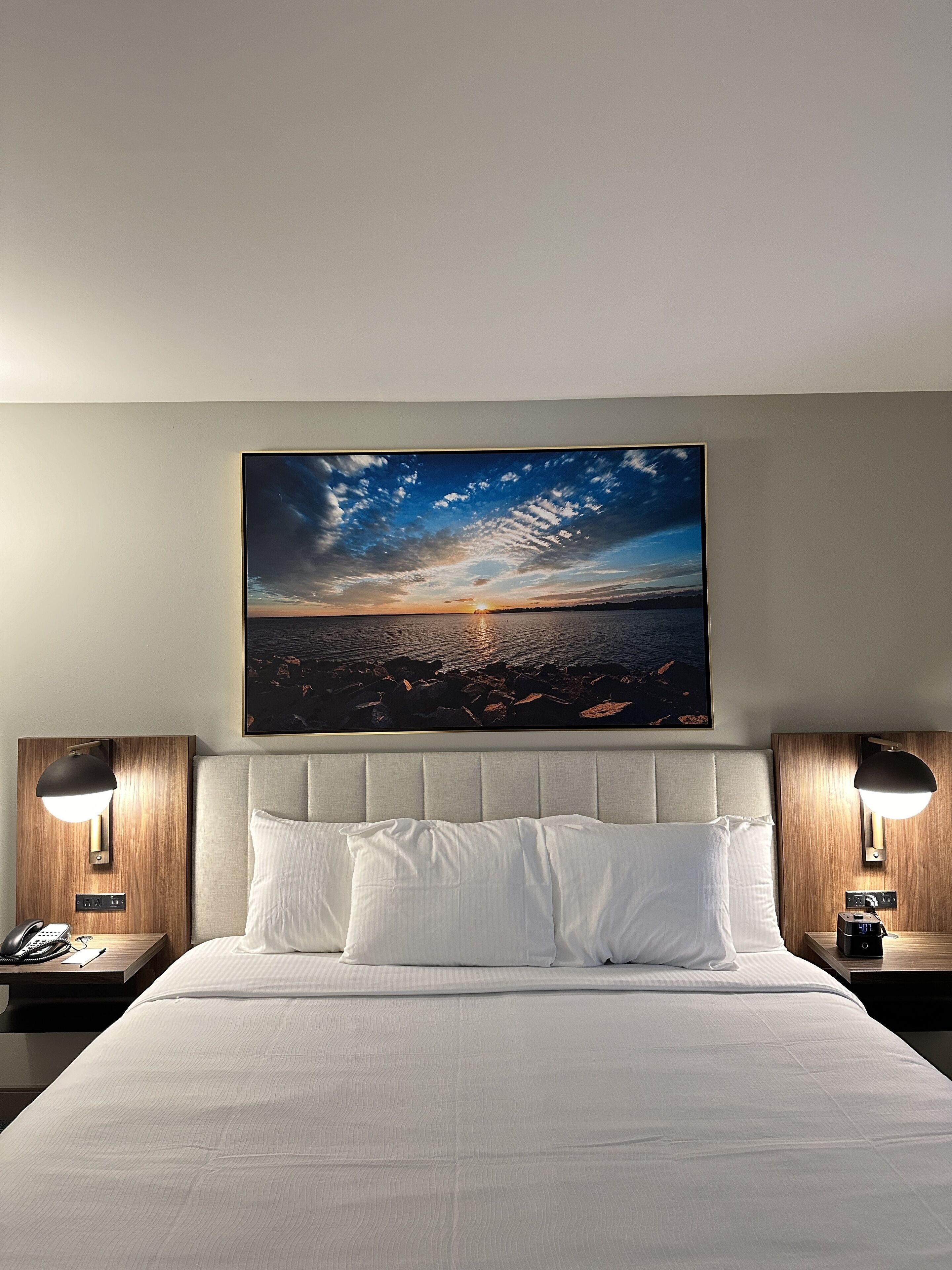 A hotel room with a large bed, two bedside tables with lamps, and a wall-mounted painting of a sunset over water.