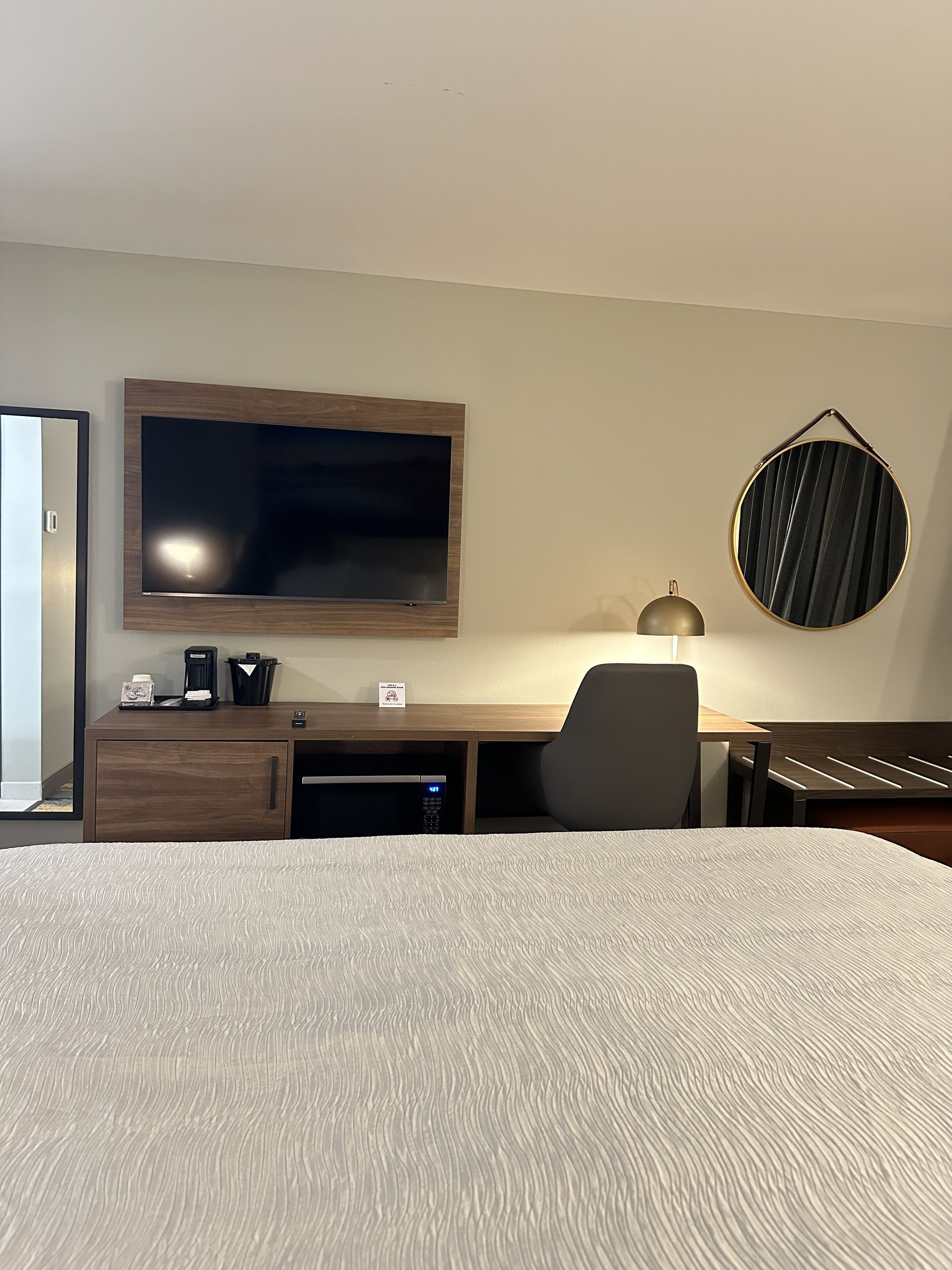 A hotel room with a large bed, a desk with a flat-screen TV, a lamp, and a circular wall decoration.