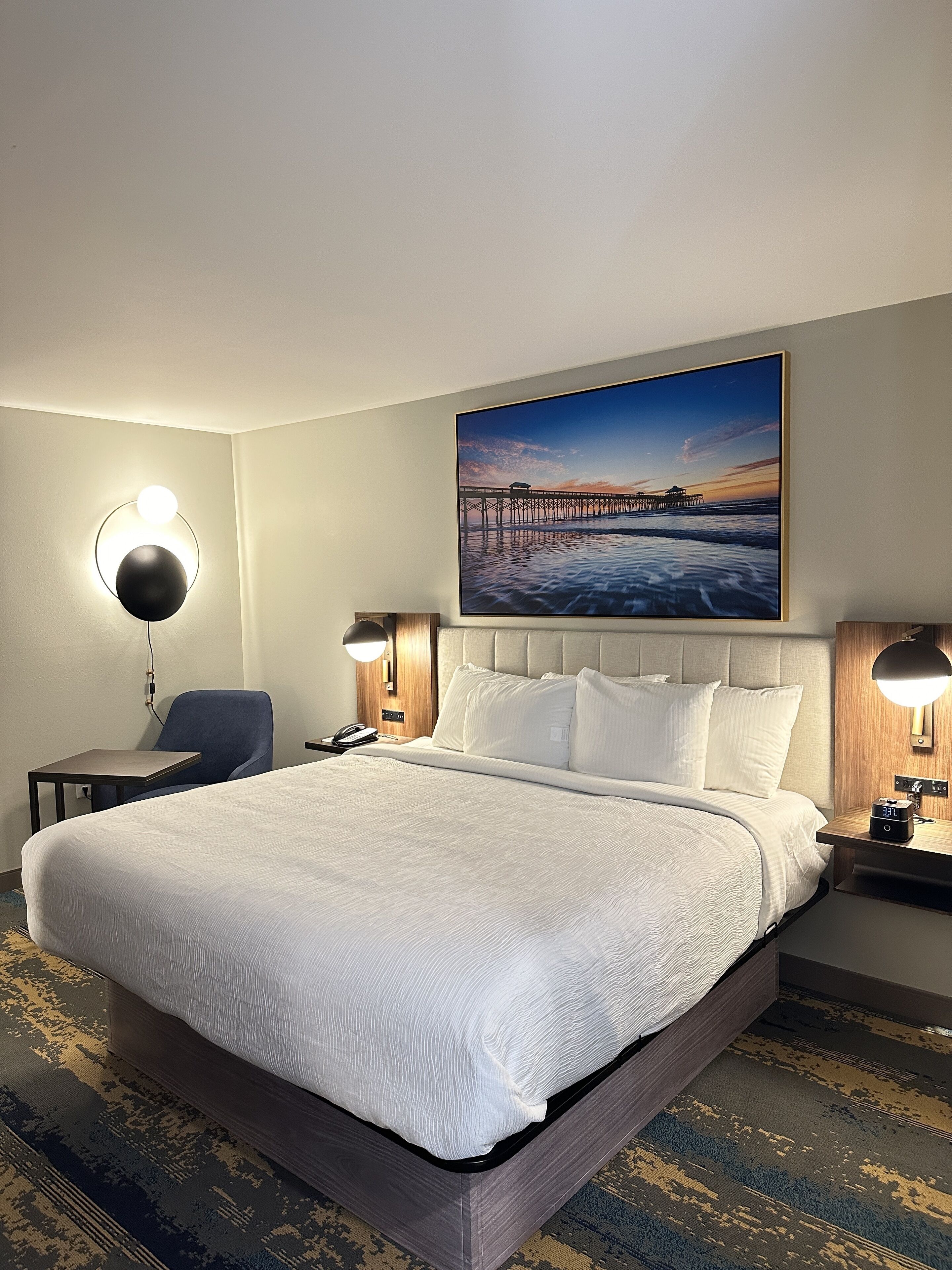 A hotel room with a large bed, a desk, a chair, and a painting of a pier on the wall.