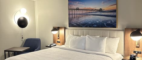A hotel room with a large bed, a desk, a chair, and a painting of a pier on the wall.