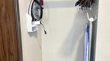 Een hoek van een kamer met een houten rek met meerdere paar schoenen, een wit strijkplank met een strijkijzer, en een blauw schoonmaakmiddel.