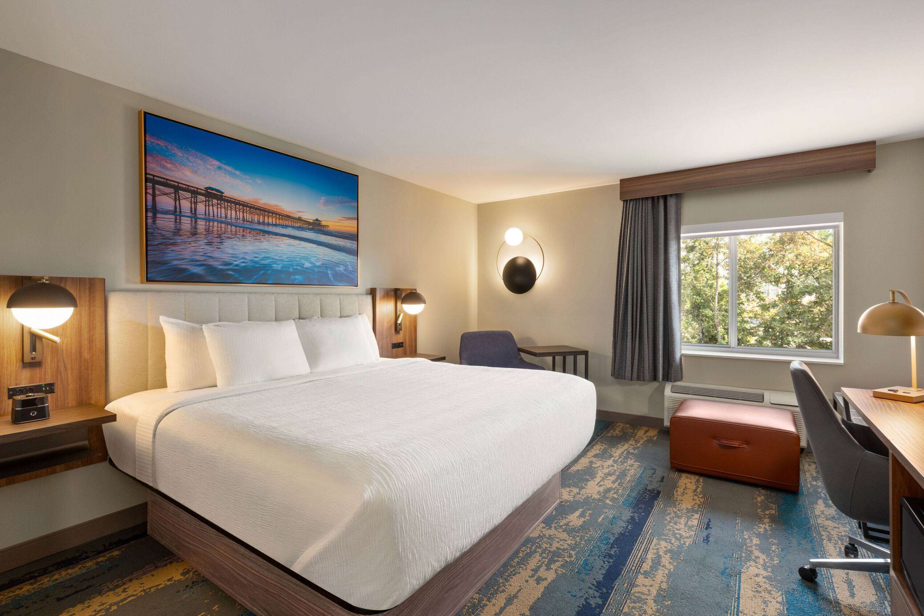 Quarto de hotel com uma cama grande, uma escrivaninha, uma cadeira, janela com vista e um quadro na parede.