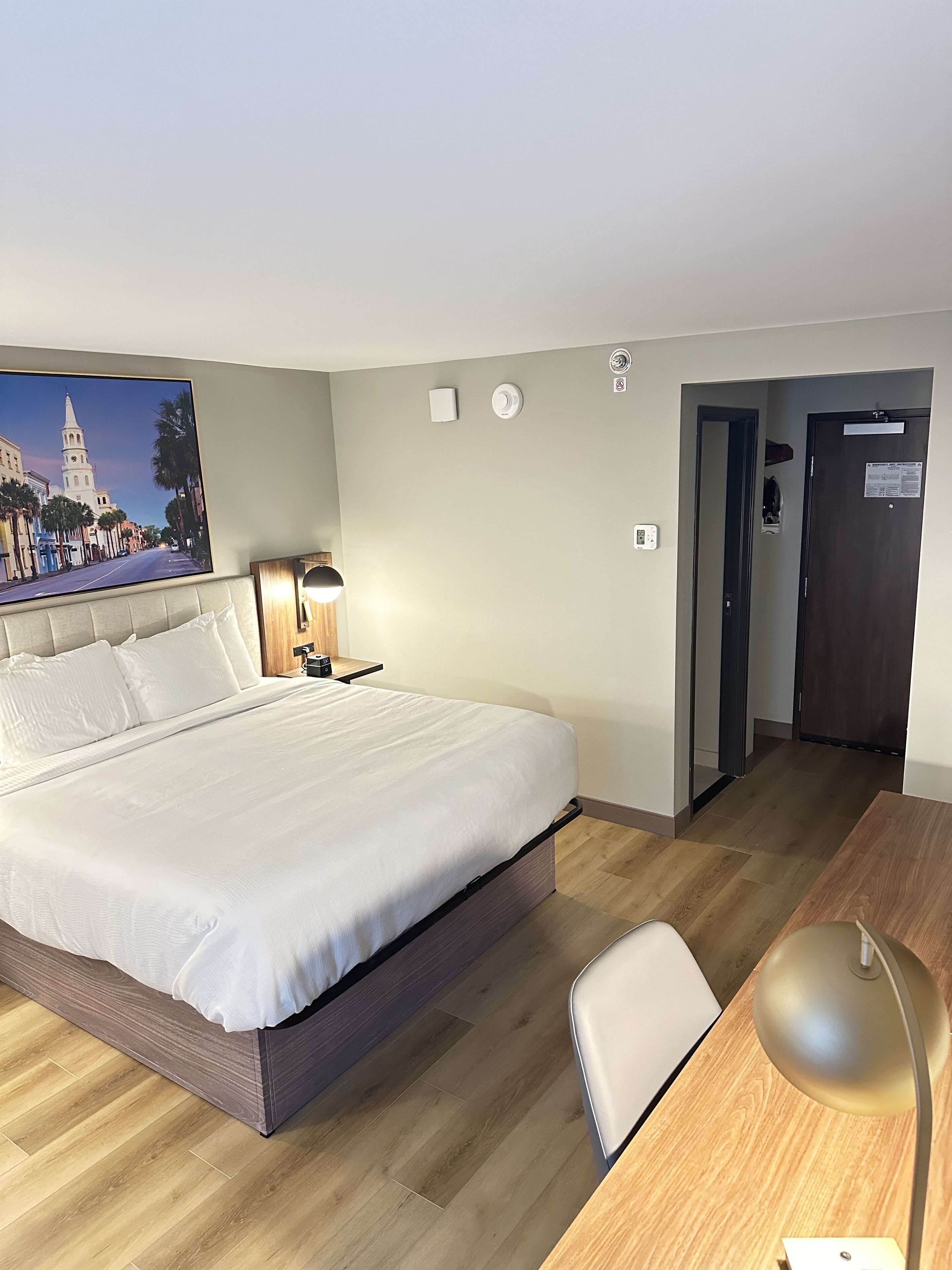 A hotel room with a large bed, a desk with a lamp, a nightstand, and a door leading to another room.