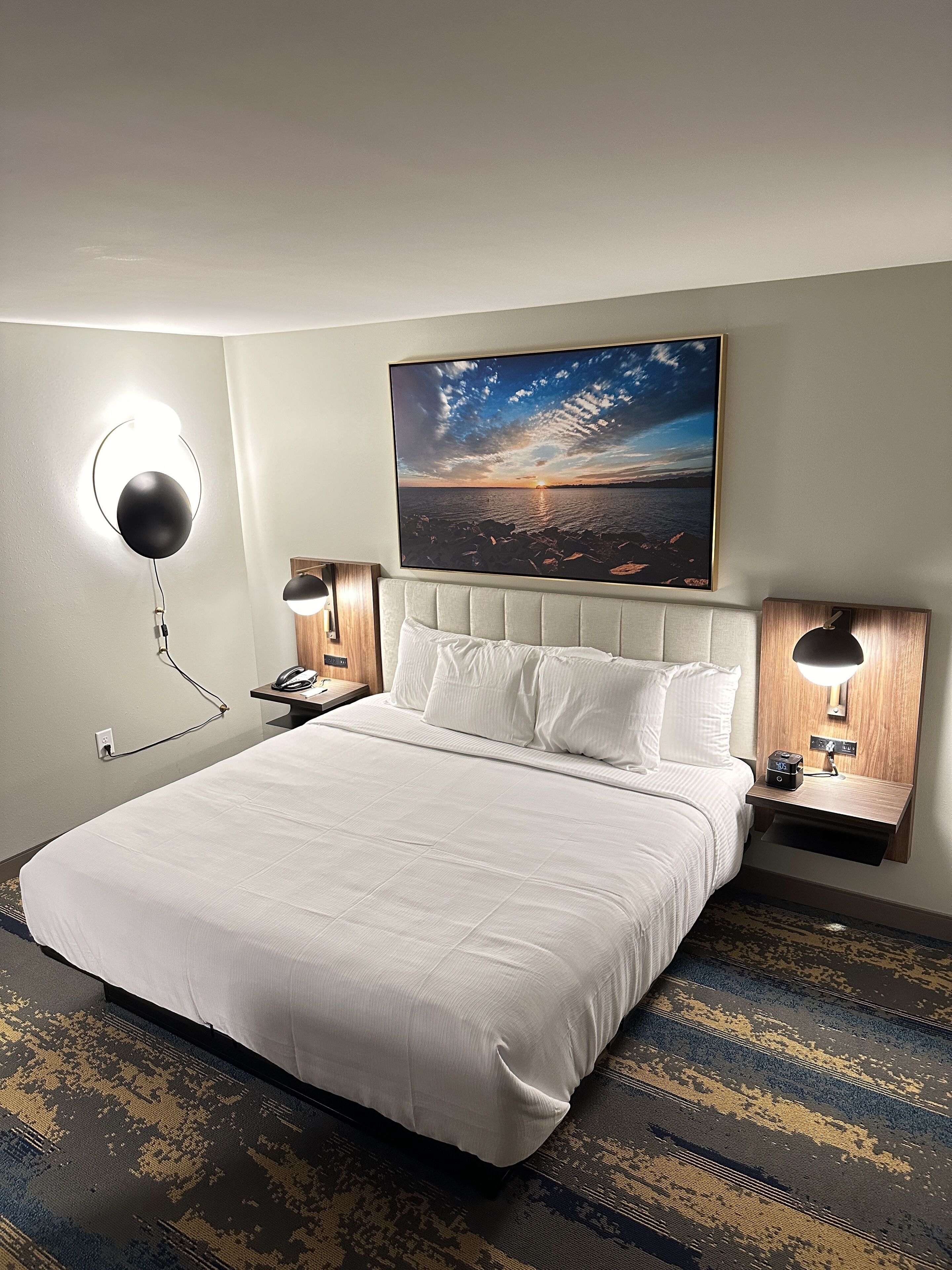 A hotel room with a large bed, two bedside tables with lamps, a wall-mounted painting, and a patterned carpet.