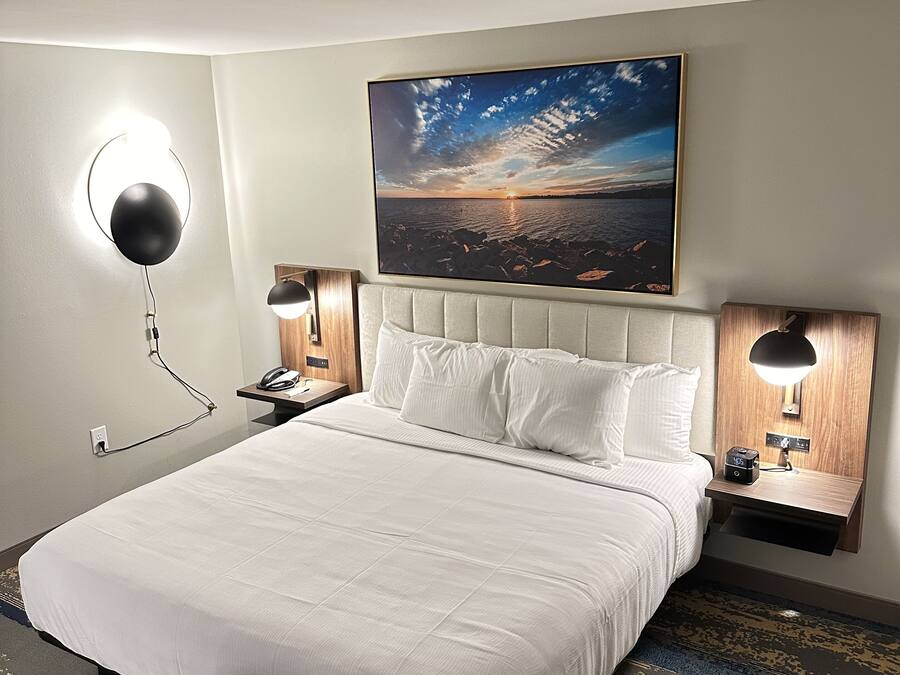 Habitación de hotel con una cama grande, dos mesitas de noche con lámparas, un cuadro colgado en la pared y una alfombra estampada.