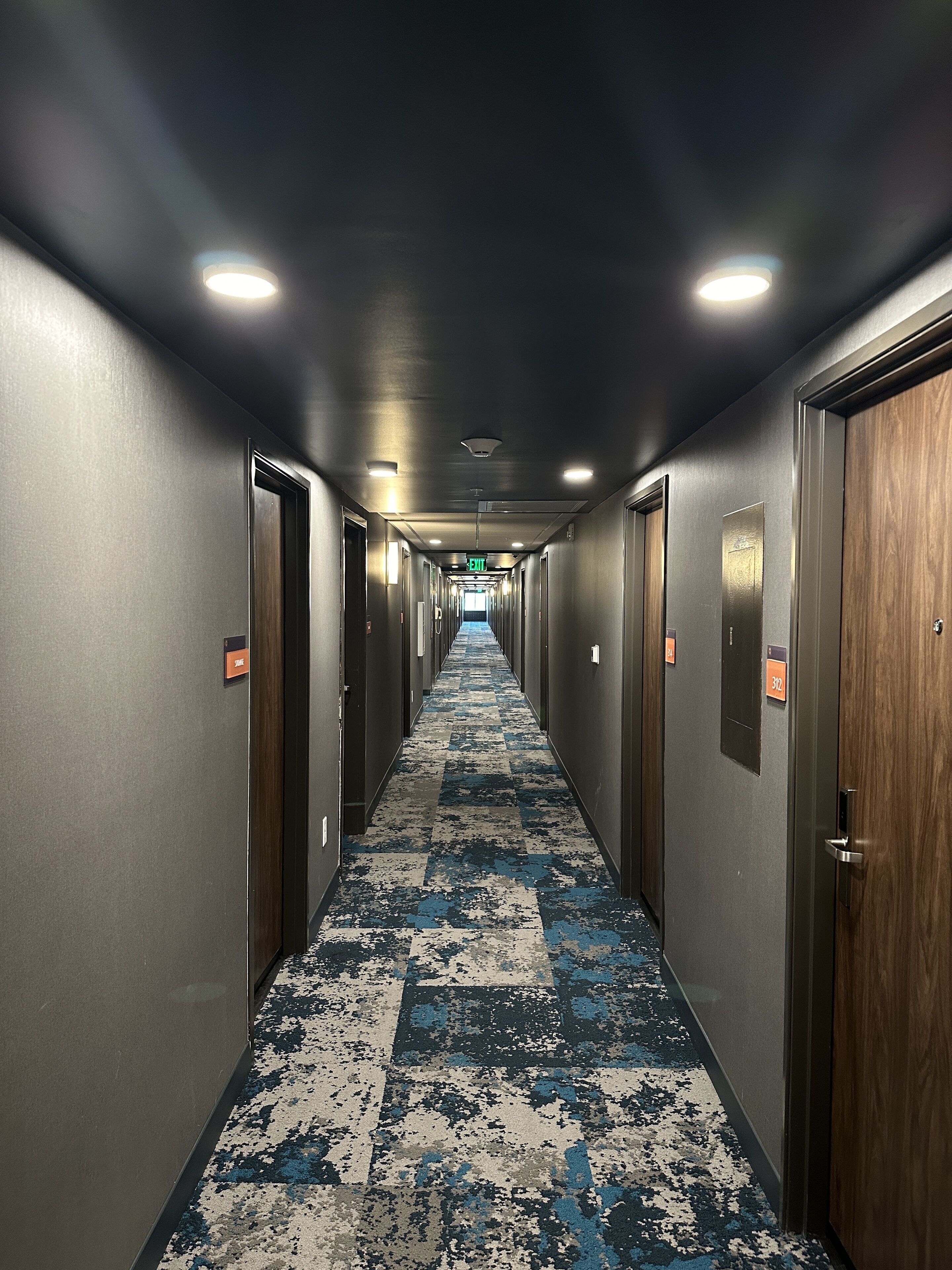 A hotel hallway with a checkered carpet, doors on both sides, and an exit sign at the end.