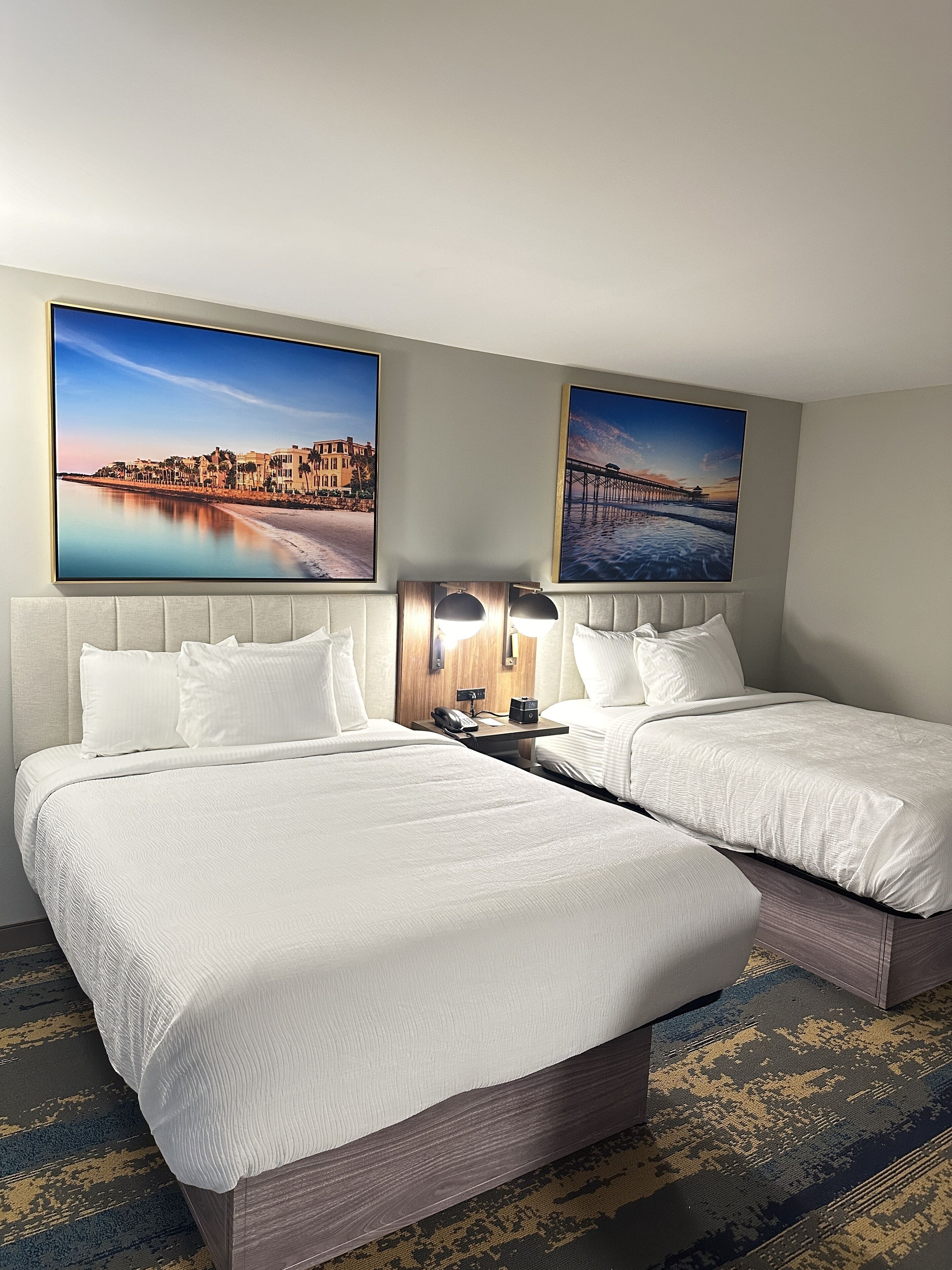 A double bed with white linens, a bedside table with a lamp, and two framed pictures on the wall.