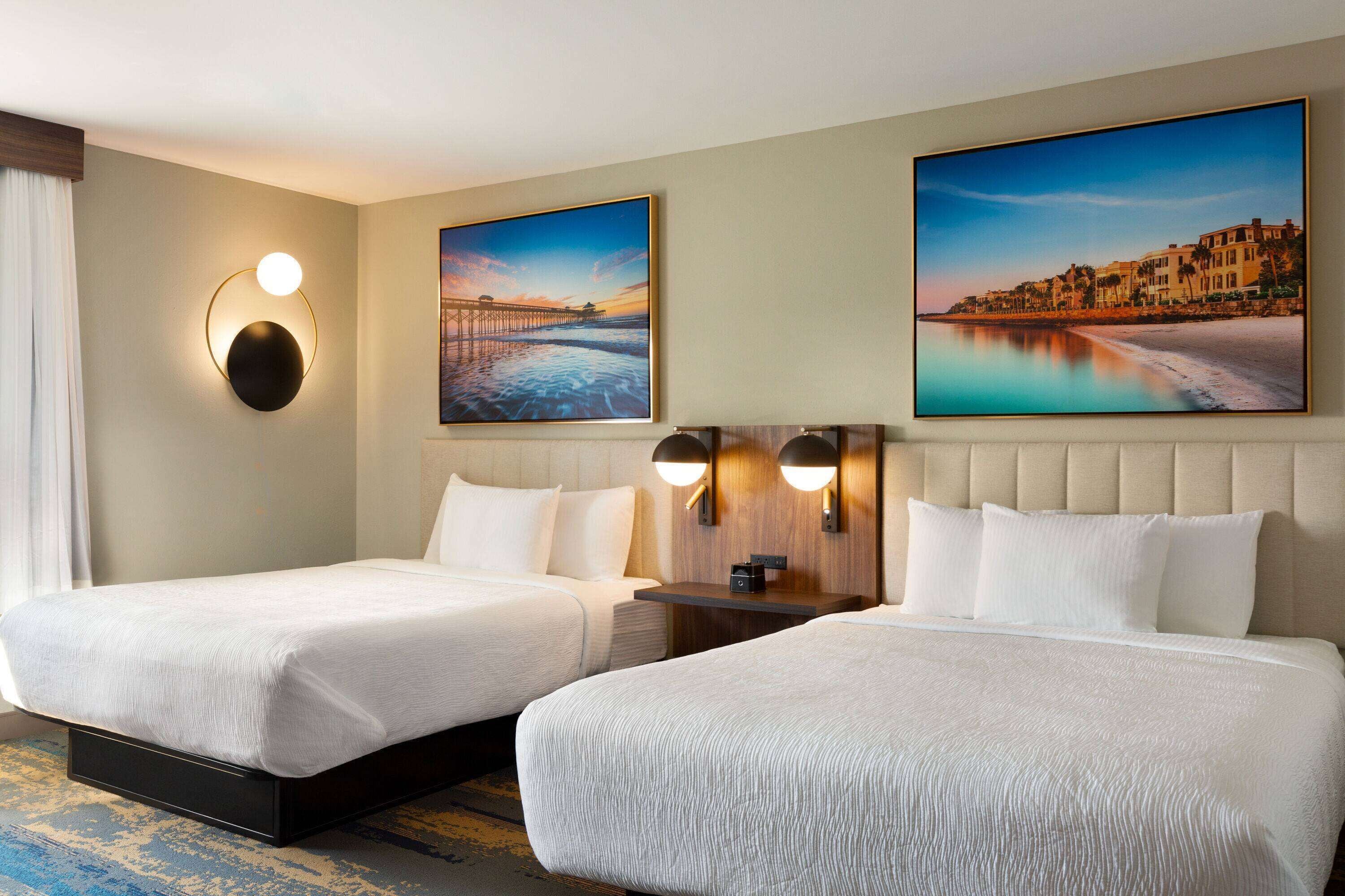 A hotel room with two beds, a large painting of a coastal scene, and a round wall light.