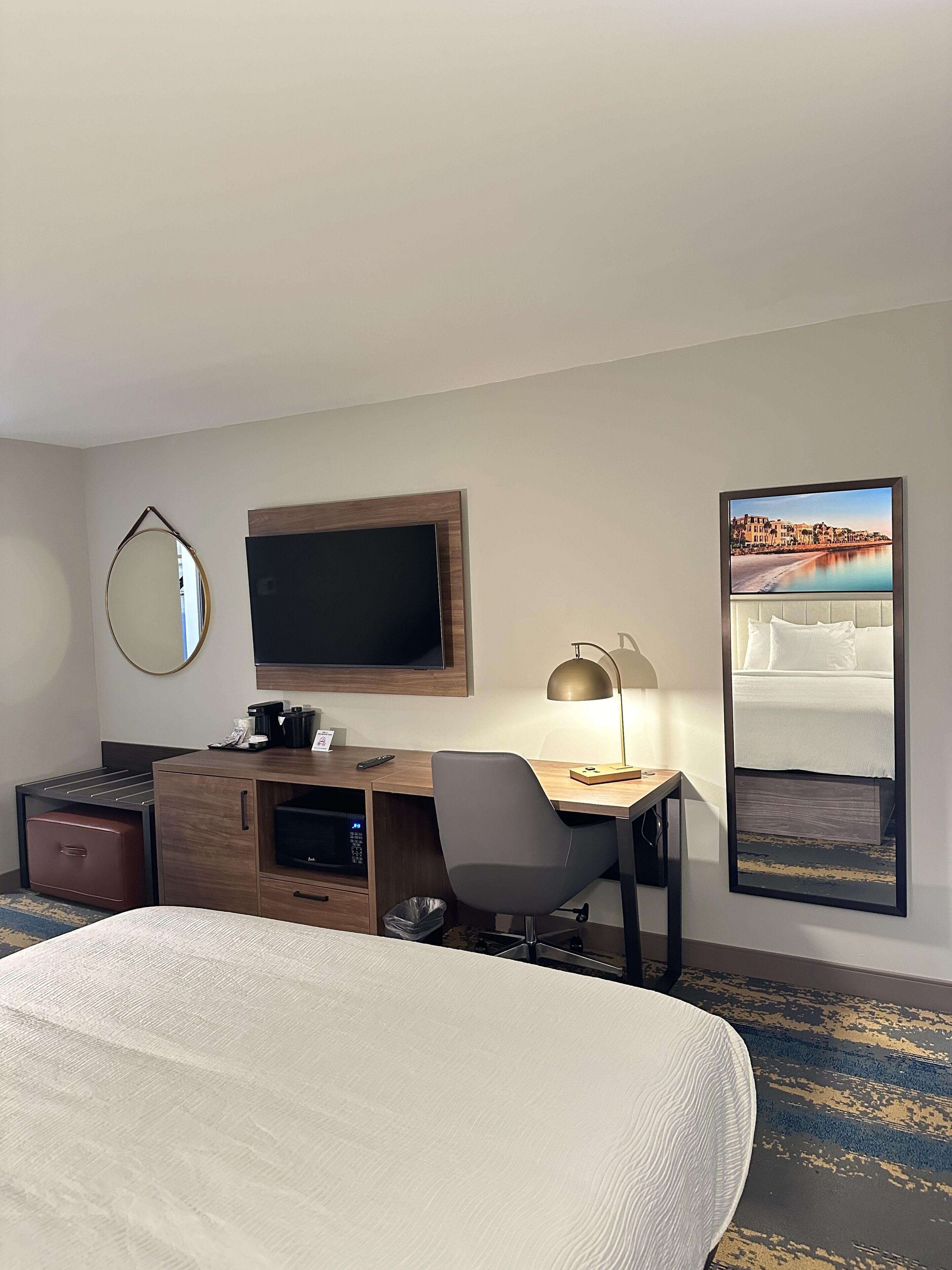 A hotel room with a bed, a desk with a computer, a TV, a mirror, and a framed picture on the wall.