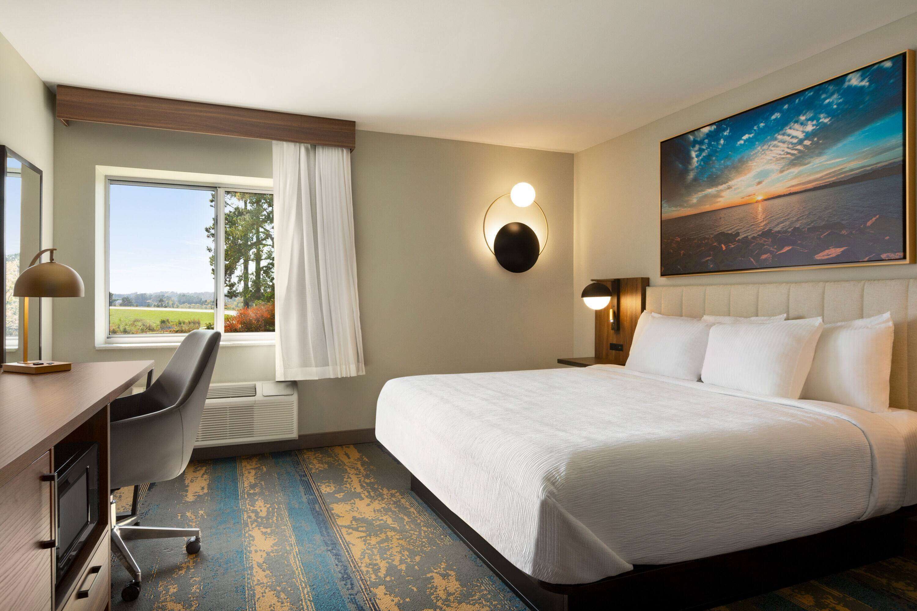 A hotel room with a large bed, a desk with a chair, a large window with curtains, a wall-mounted light, and a painting of a sunset.