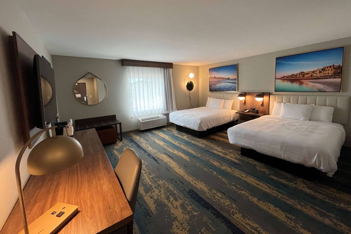 A hotel room with two beds, a desk with a chair, a desk lamp, a mirror, a window with curtains, an air conditioning unit, and two wall-mounted lamps.