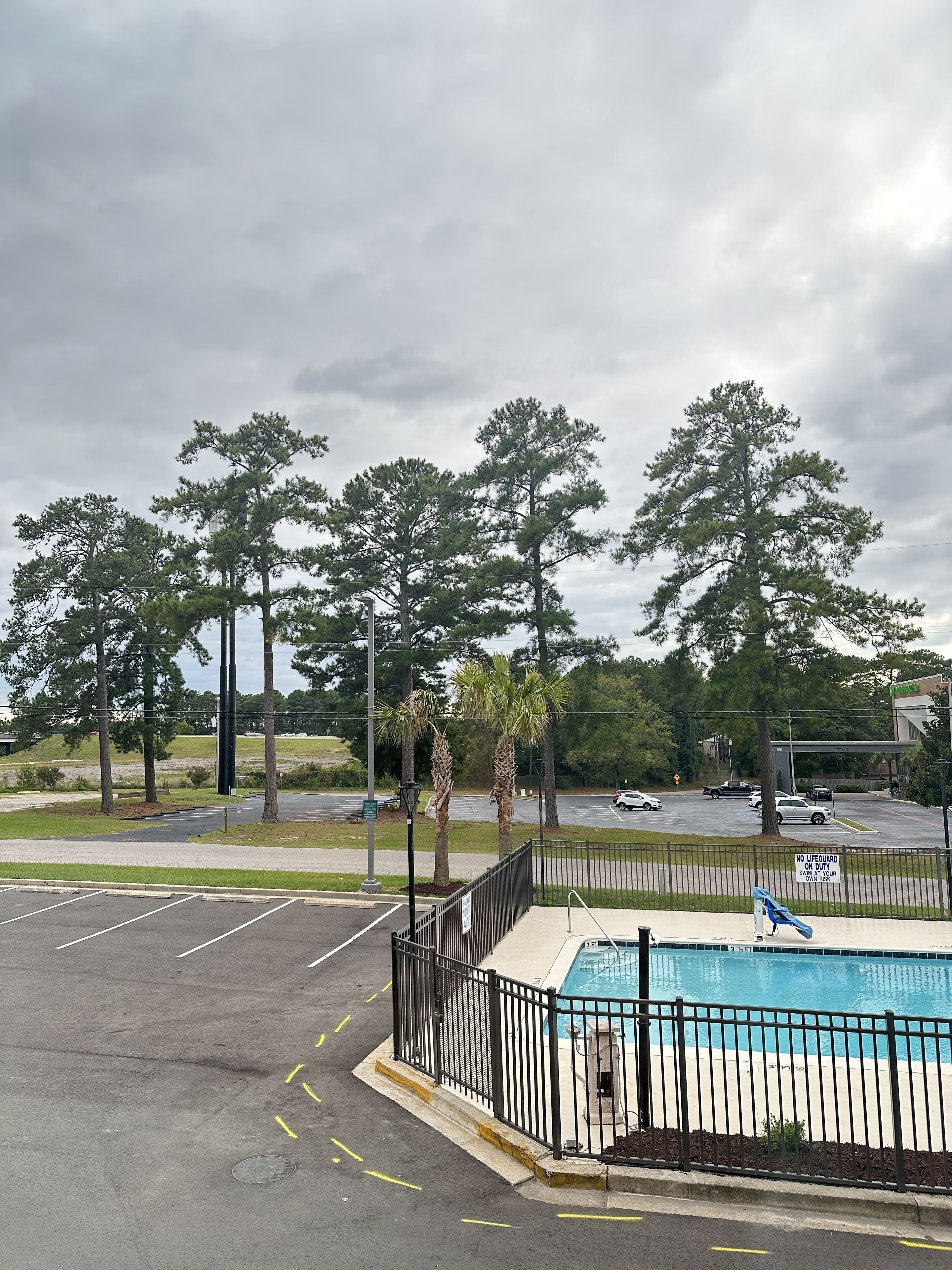 A parking lot with a swimming pool, surrounded by trees and a road with vehicles.