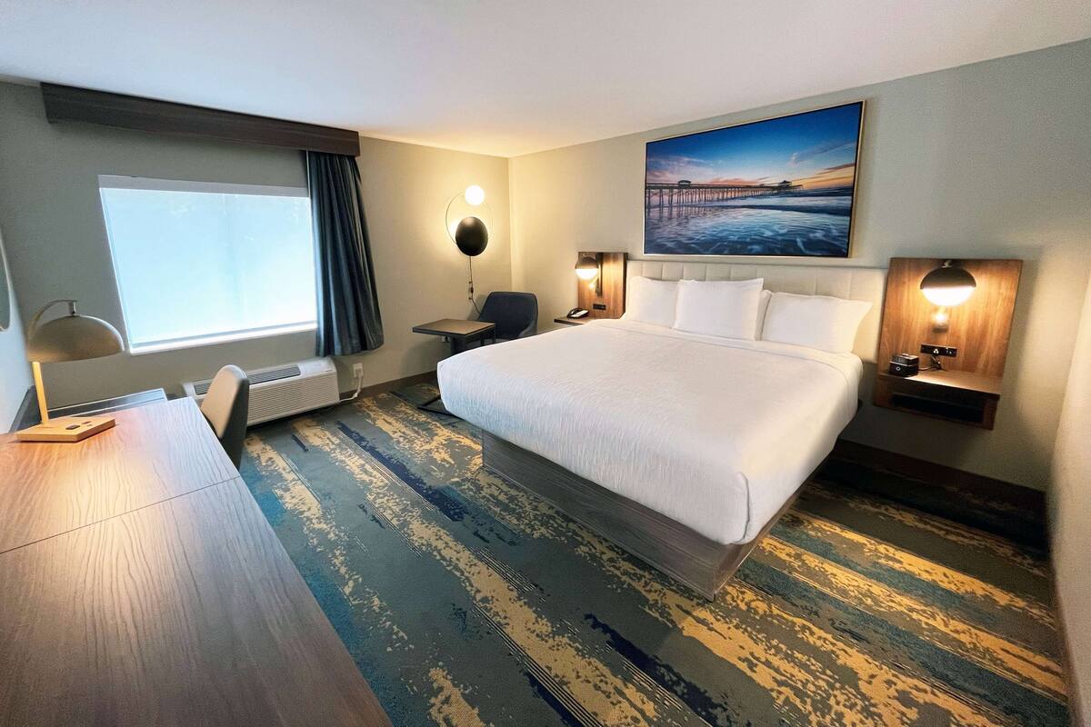 A hotel room with a large bed, a desk, a chair, a window with curtains, a painting on the wall, and a carpeted floor.