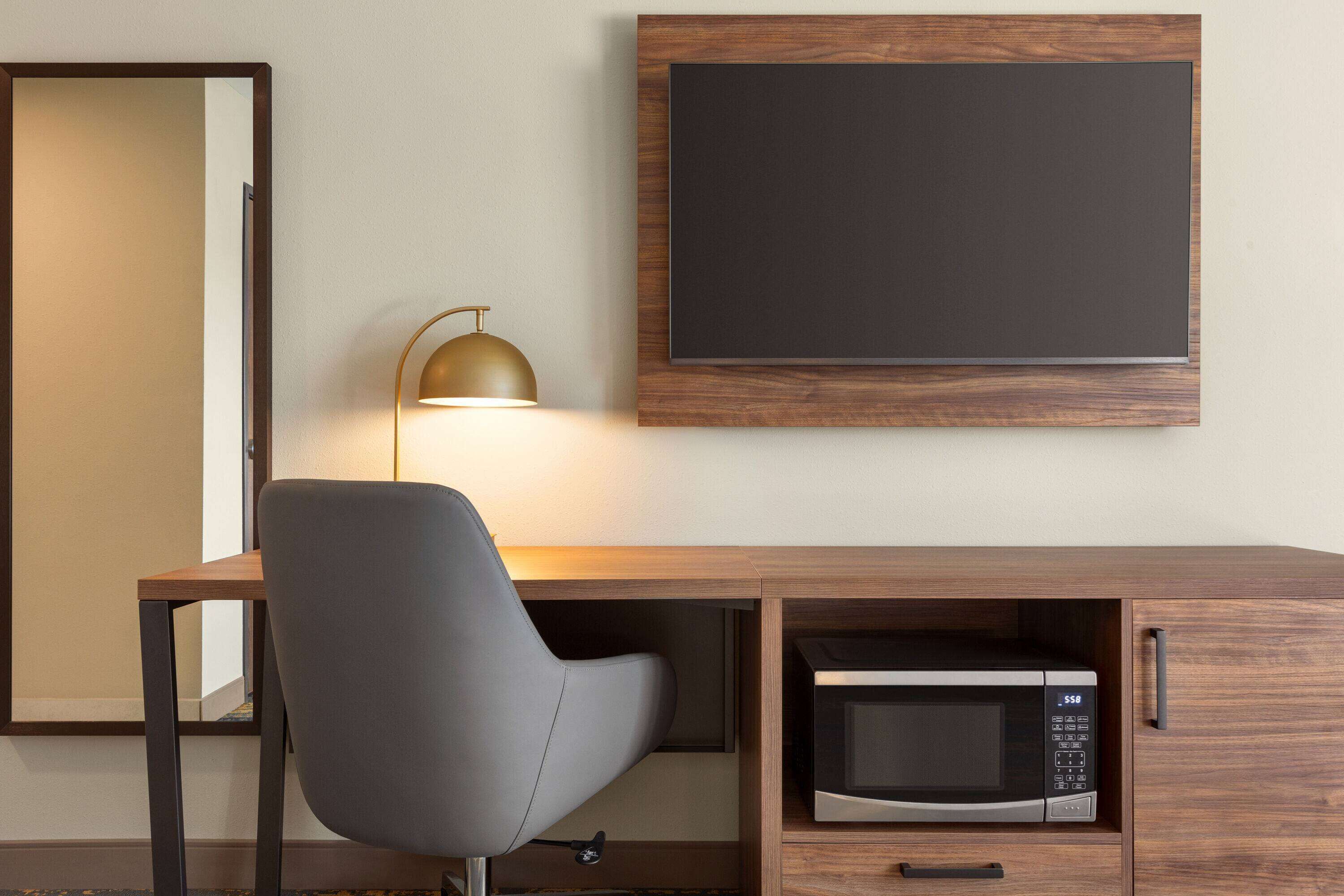 Habitación de hotel moderna con un escritorio de madera, una silla gris, un televisor montado en la pared y un microondas integrado.