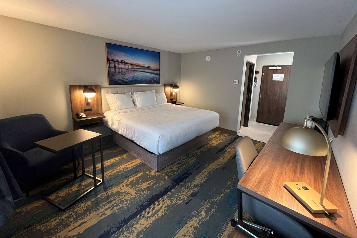 A hotel room with a large bed, bedside tables with lamps, a desk with a chair, a TV, and a painting on the wall.