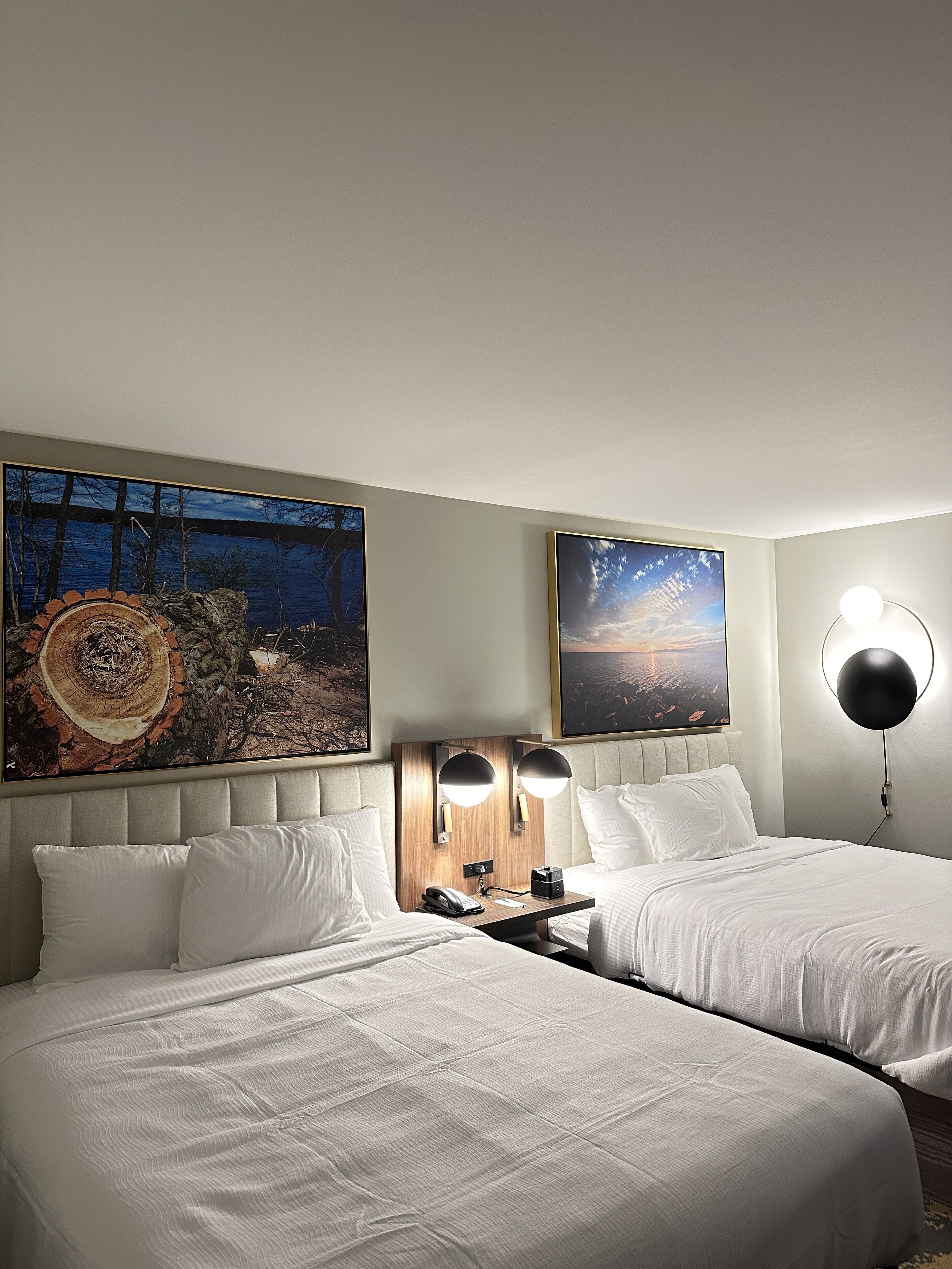 A hotel room with two beds, a wooden headboard, a nightstand with a lamp, and two large paintings on the wall.