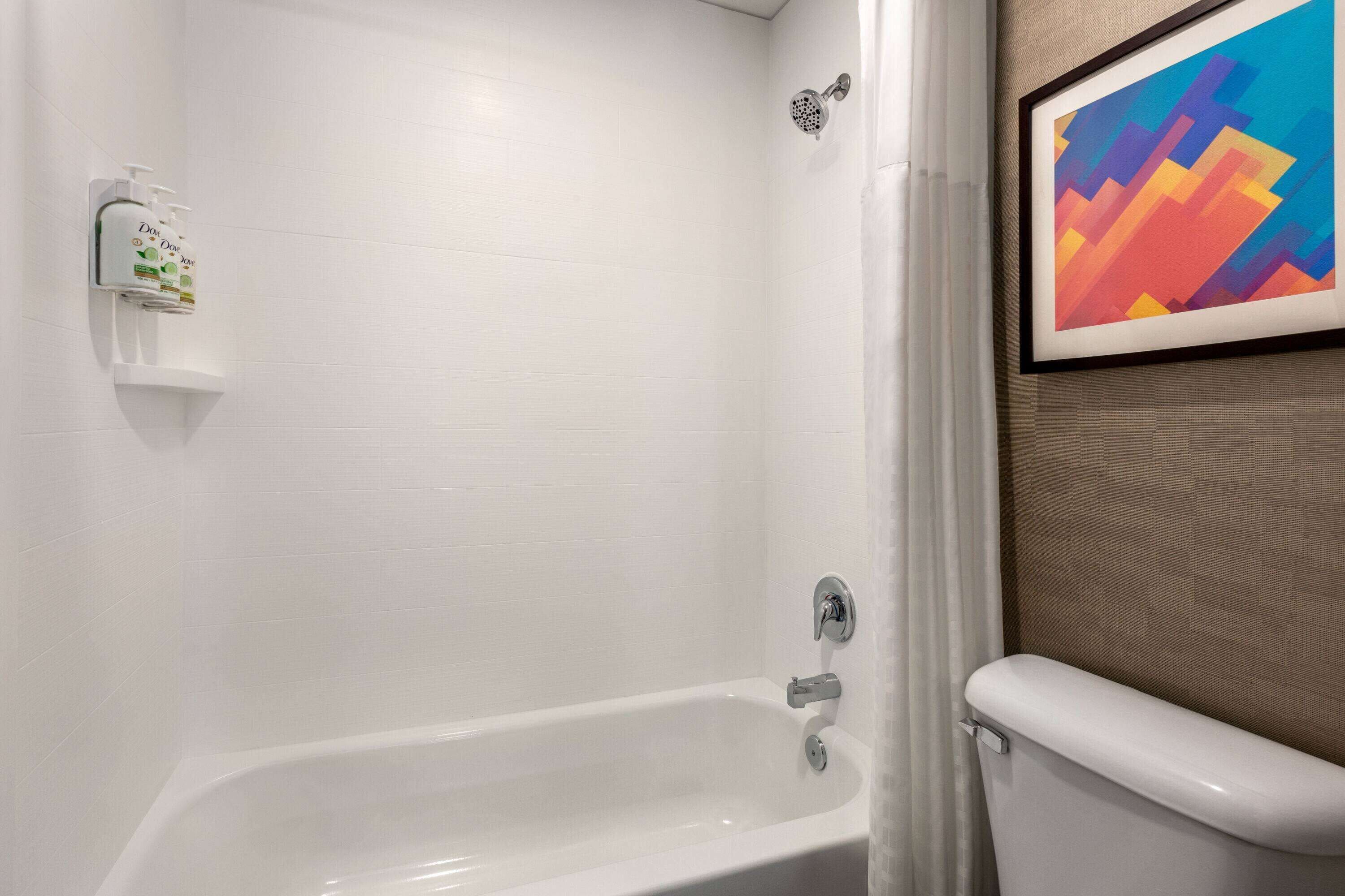 A bathroom with a white bathtub, a showerhead, and a toilet. There is a framed abstract painting on the wall.