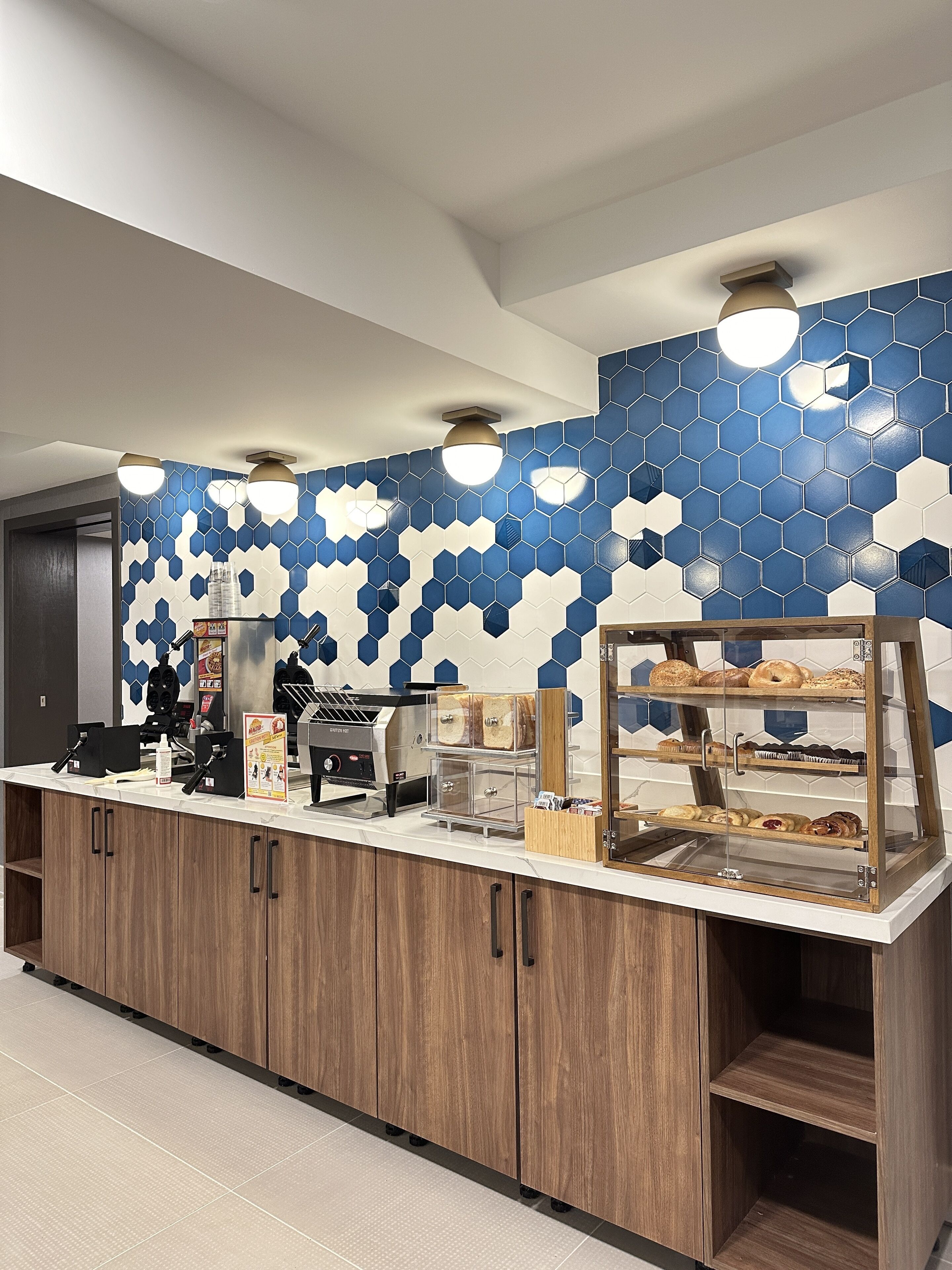Un comptoir pour le petit-déjeuner avec une machine à café, une vitrine remplie de pâtisseries et un mur orné d’un motif hexagonal bleu et blanc.
