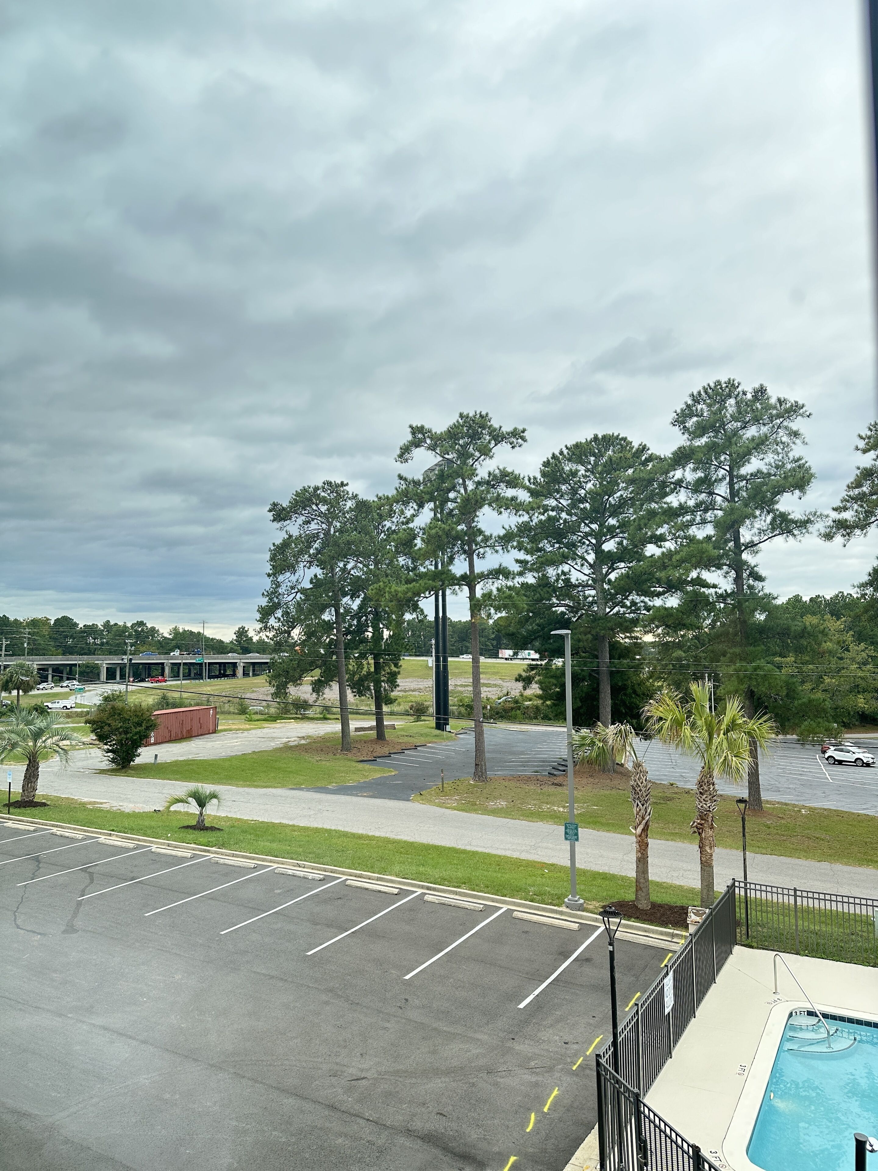 A parking lot with marked spaces, a swimming pool, and a view of a building and trees in the background.