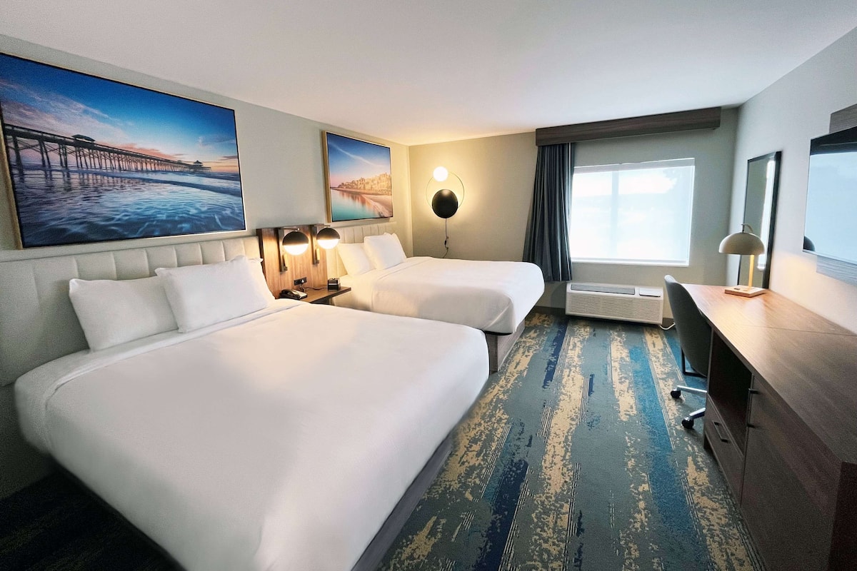 A hotel room with two beds, a desk, a chair, a large window, and artwork on the walls.
