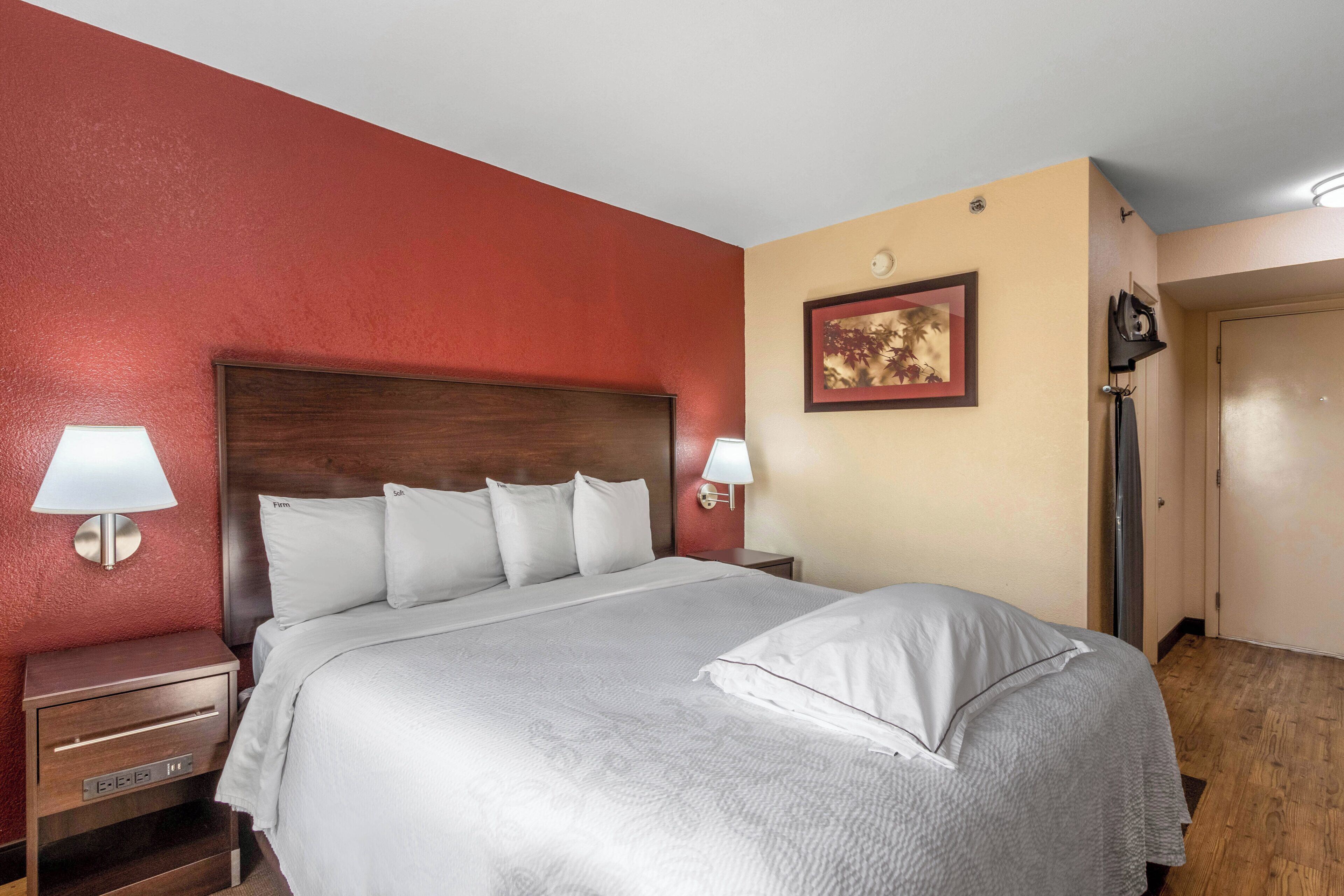A hotel room with a large bed, two bedside lamps, a nightstand, a wooden headboard, a framed picture on the wall, and a door.