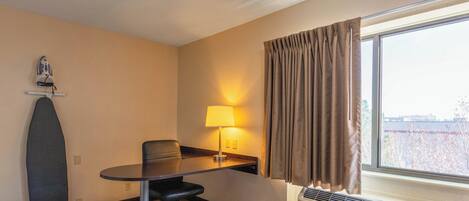 Habitación de hotel con piso de madera, un escritorio con silla, una lámpara, una ventana con cortinas y un equipo de aire acondicionado.