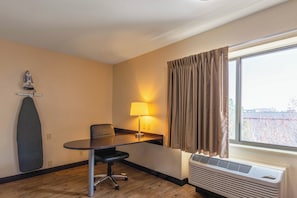 A hotel room with a wooden floor, a desk with a chair, a lamp, a window with curtains, and an air conditioning unit. - Red Roof Inn PLUS+ El Paso East (El Paso)