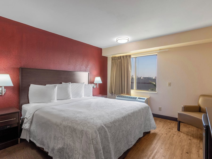 Una habitación de hotel con una cama grande, dos lámparas de noche, un ventanal con cortinas, una silla y una lámpara de techo.