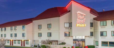 A Red Roof Plus+ hotel with a modern design, featuring a prominent neon sign, multiple windows, and a well-maintained exterior.