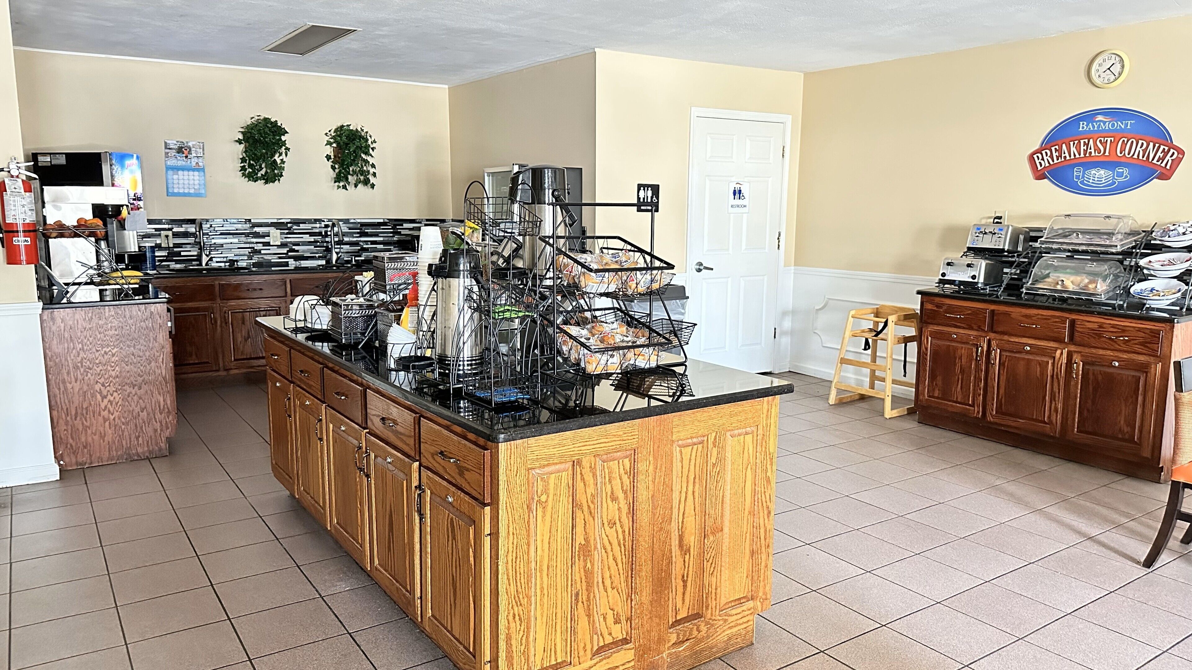 A breakfast bar with a coffee machine, toaster, and food display. There is a restroom sign and a Baymont Breakfast Corner logo on the wall.