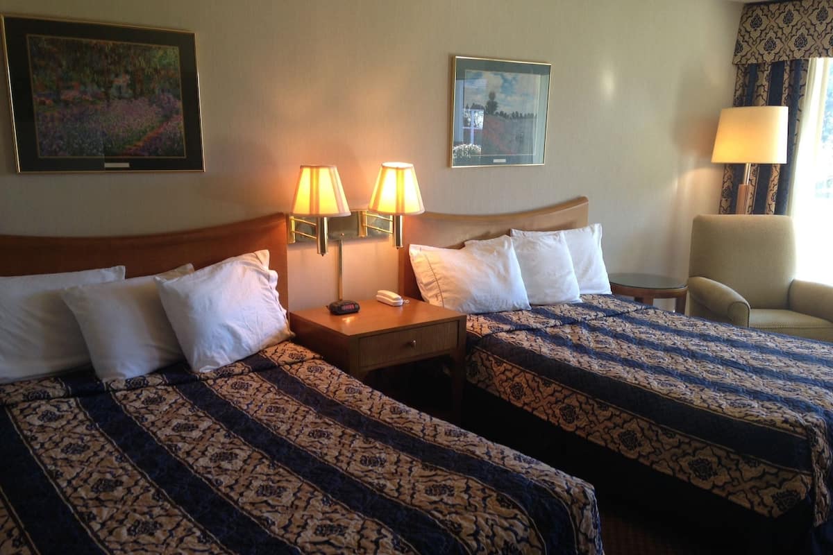 A hotel room with two beds, each with patterned bedding, a nightstand with a lamp, a chair, and artwork on the walls.