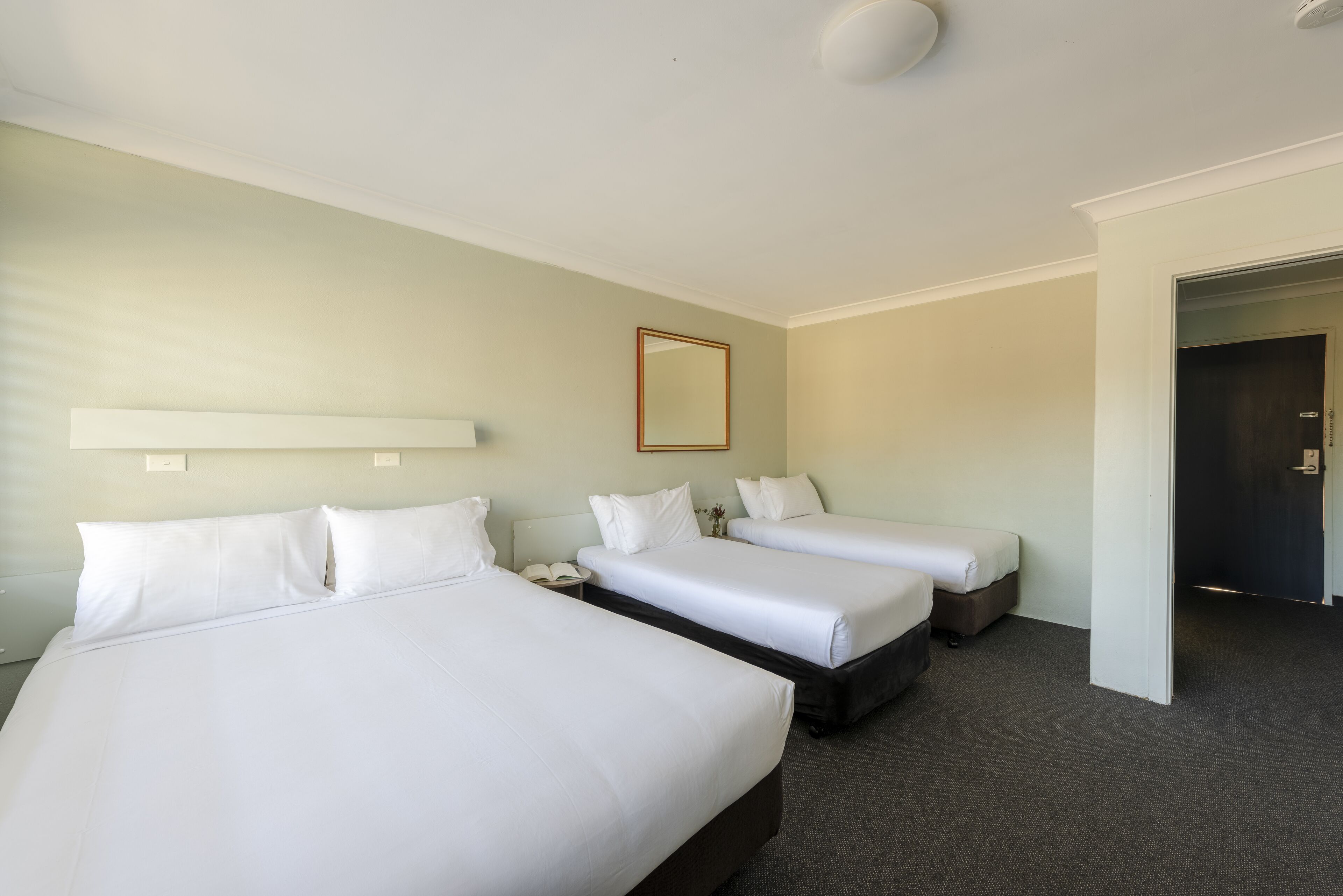A hotel room with three single beds, a bedside table with a lamp, a large mirror on the wall, and a door leading to another room.