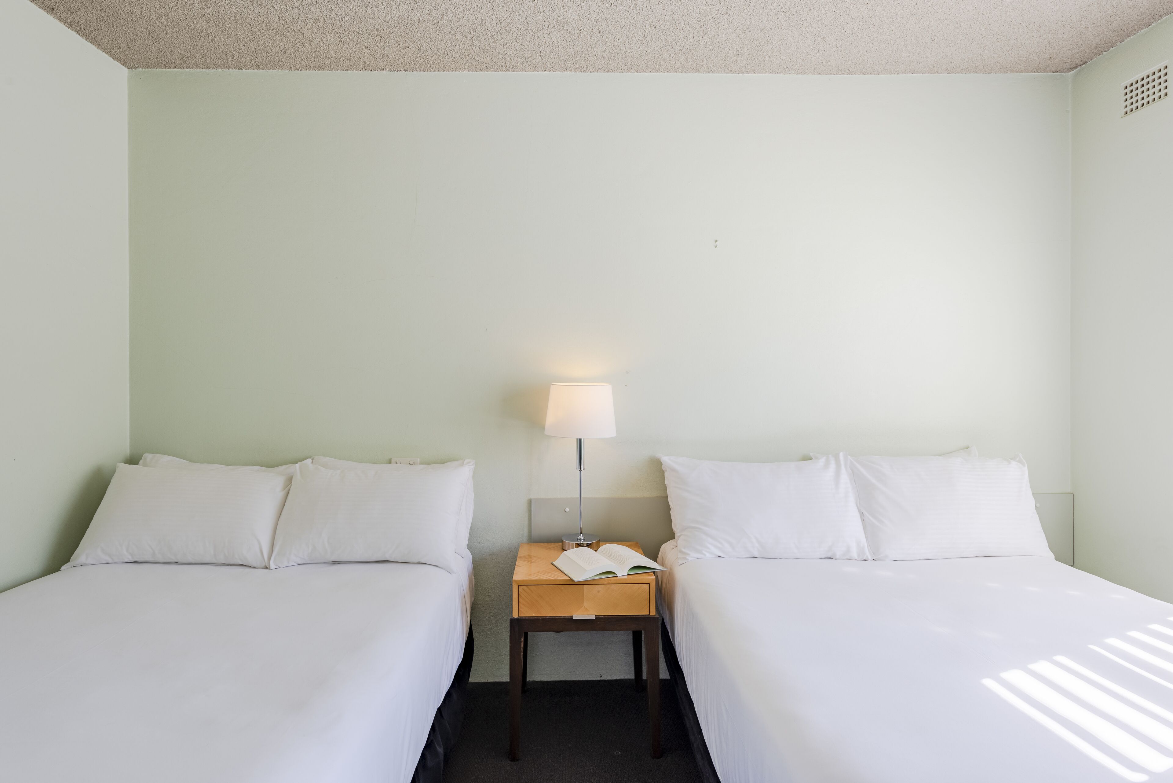 A hotel room with two single beds, a nightstand with a lamp, and a book on the nightstand.
