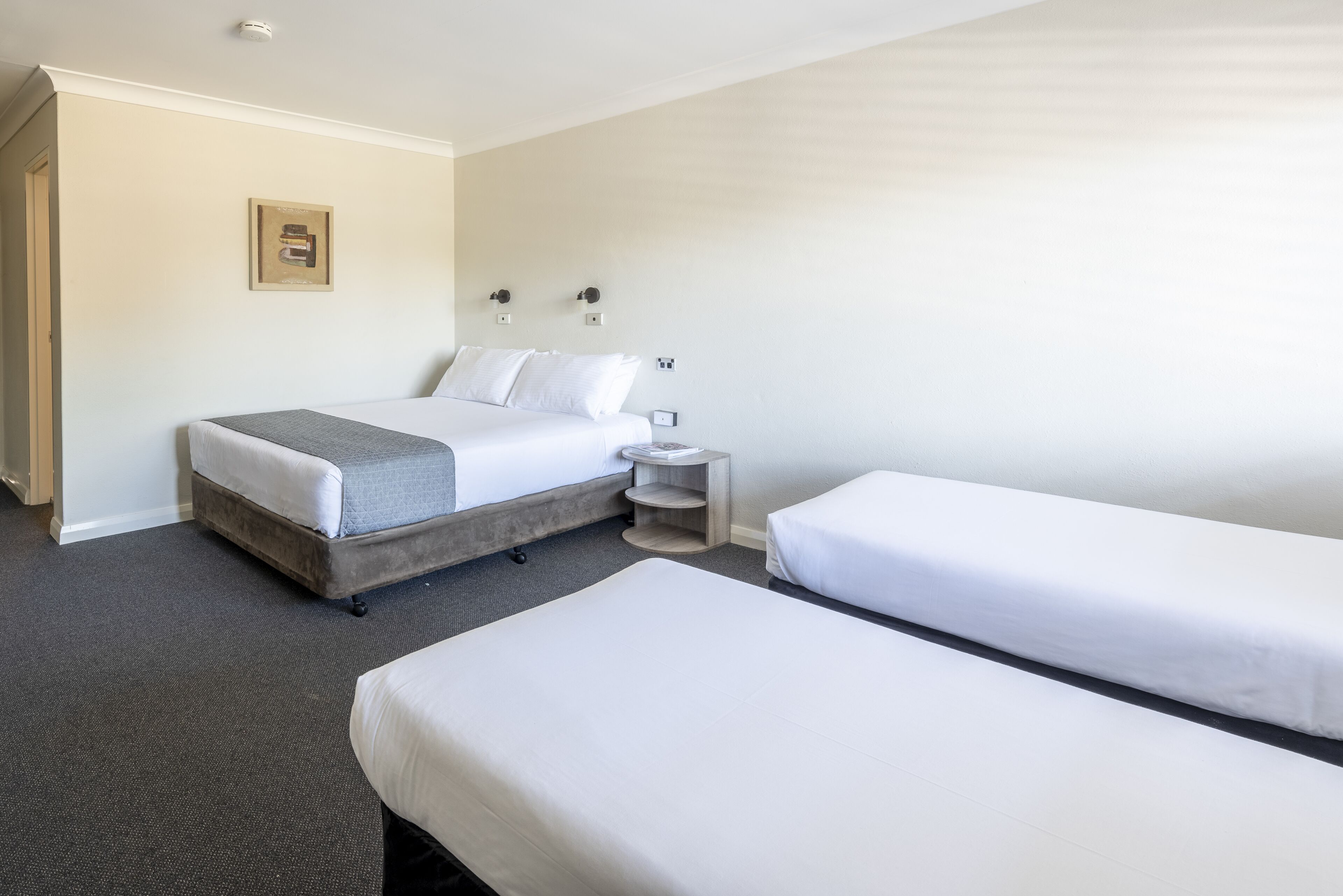 Een hotelkamer met een eenpersoonsbed, een tweepersoonsbed, een klein nachtkastje en een wandplank.