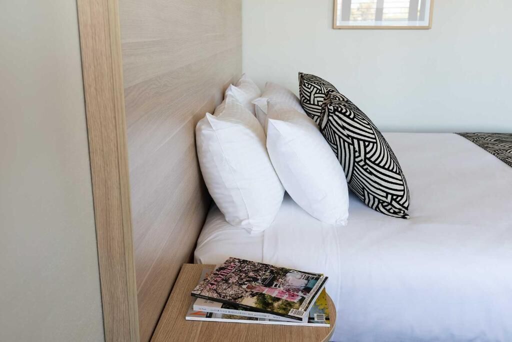 A neatly made bed with white and patterned pillows, a magazine on a bedside table, and a framed picture on the wall.