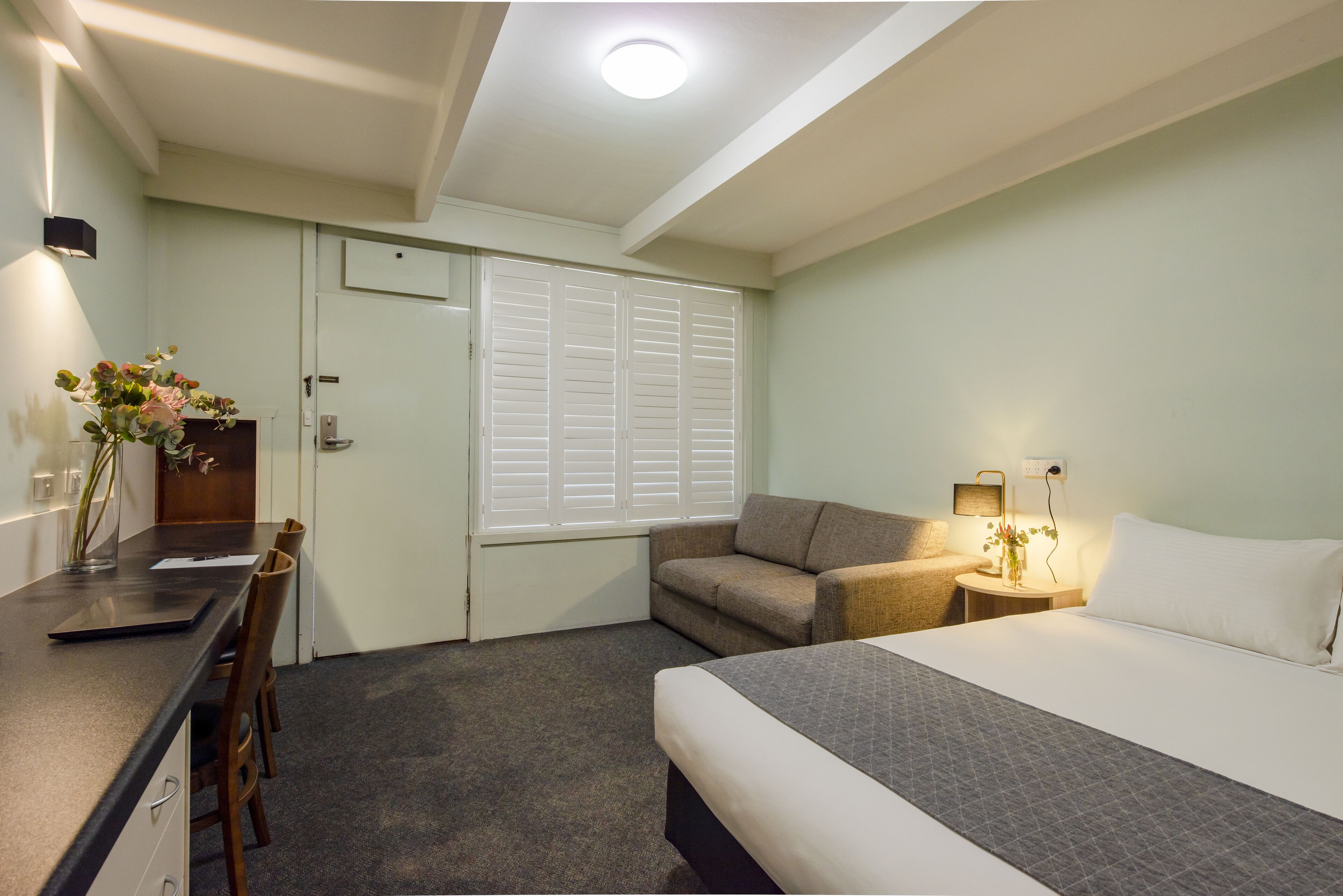 A hotel room with a bed, a desk with a chair, a sofa, a bedside table with a lamp, and a window with blinds.
