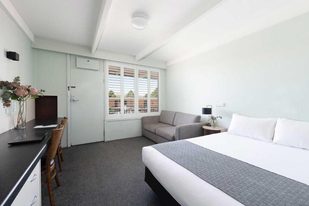 Een hotelkamer met een bed, een bureau, een stoel, een bank, een lamp en een raam met jaloezieën.
