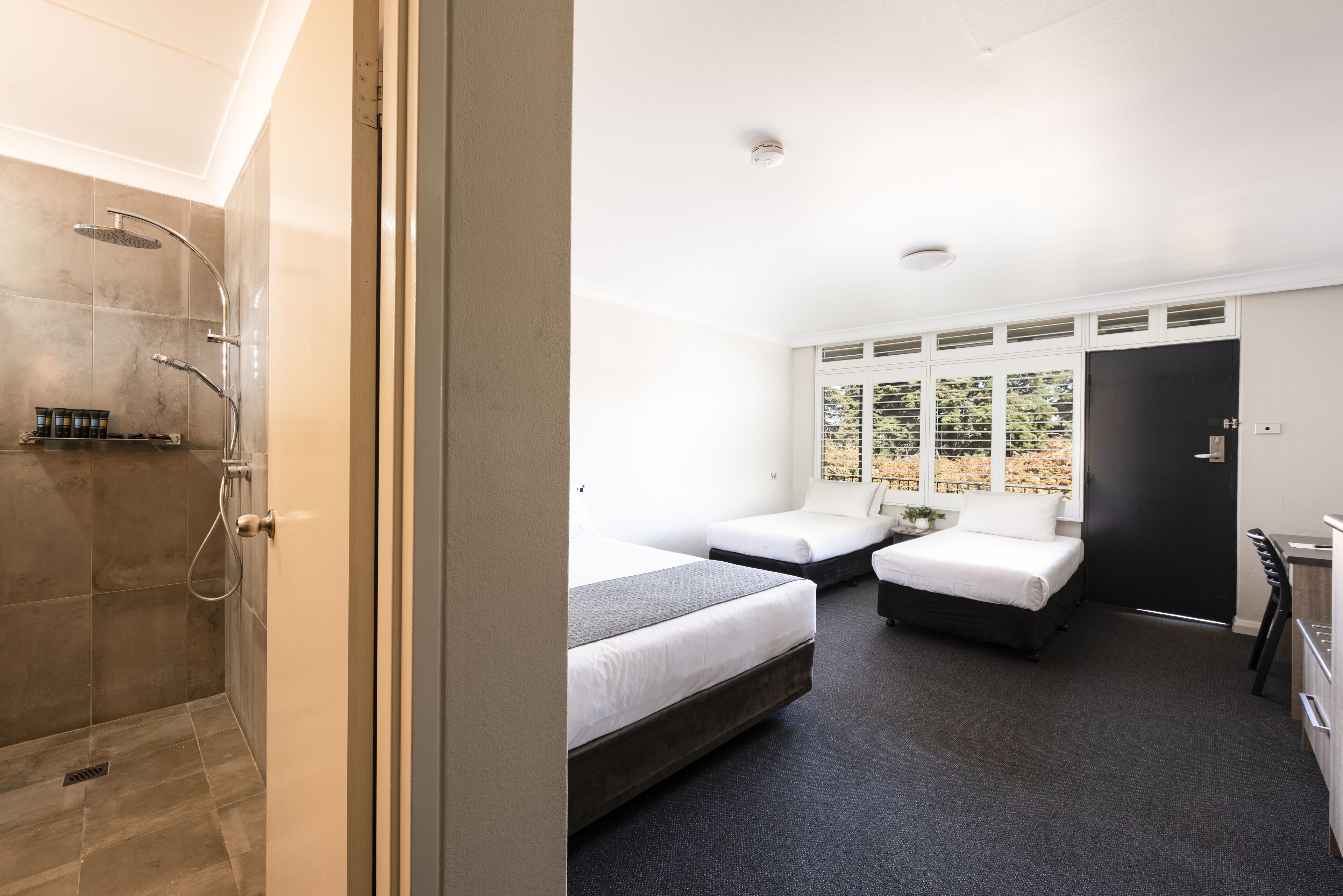 A hotel room with a shower, two beds, a desk, and a window with a view of trees.