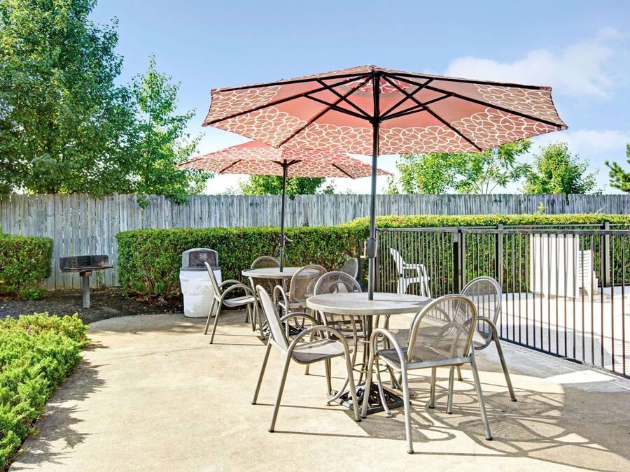Een terras buiten met metalen stoelen en tafels, een groot rood parasol en een houten hek op de achtergrond.
