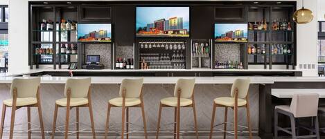 A modern bar area with a long counter, multiple seating options, and a large TV screen displaying a cityscape.