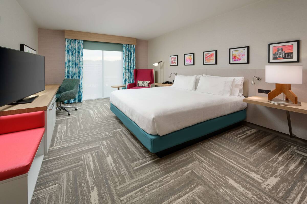 Una habitación de hotel con una cama grande, un escritorio con televisor de pantalla plana, una silla roja y una ventana con cortinas azules.