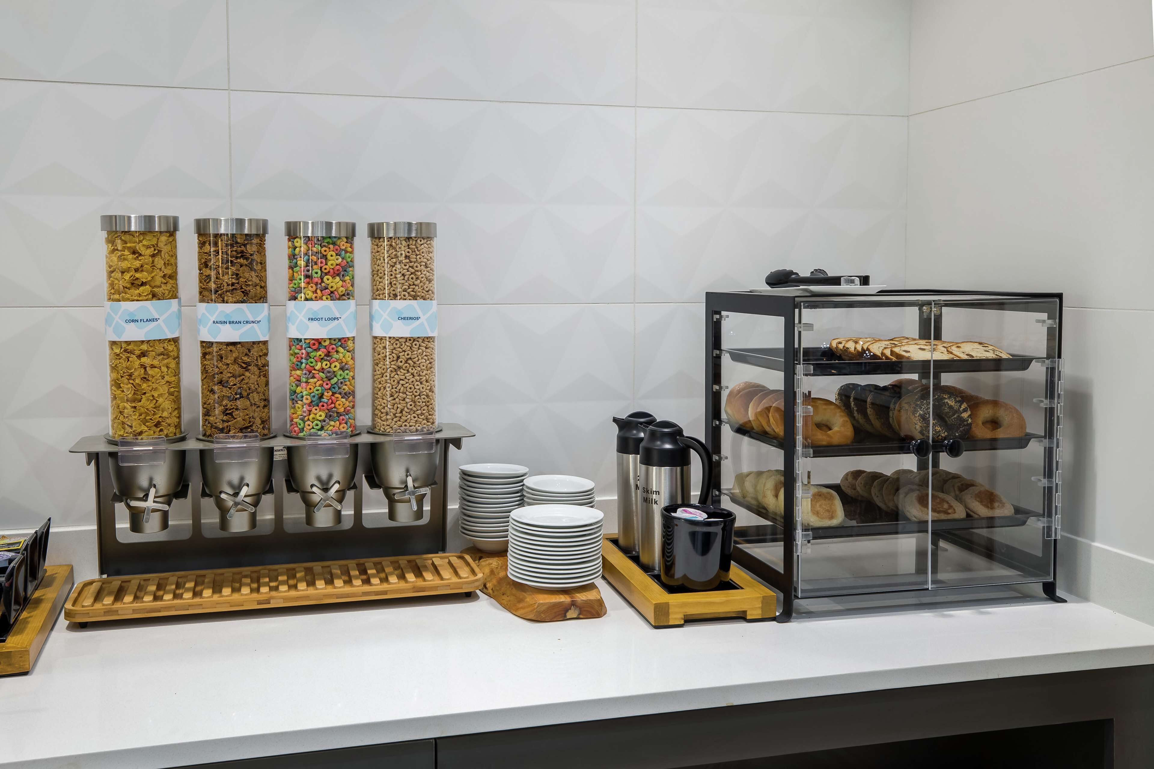A breakfast station with cereal dispensers, a coffee machine, and a glass display case with pastries.