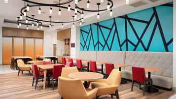 Ein modernes Restaurant mit runden Tischen, roten Stühlen, einem geometrischen Wandbild und einer gepolsterten Bank.
