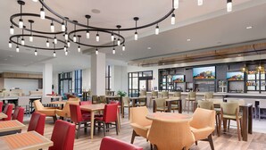 A modern restaurant with a bar area, featuring a mix of red and beige chairs, wooden tables, and pendant lighting. - Hilton Garden Inn Knoxville Papermill Drive (Knoxville)