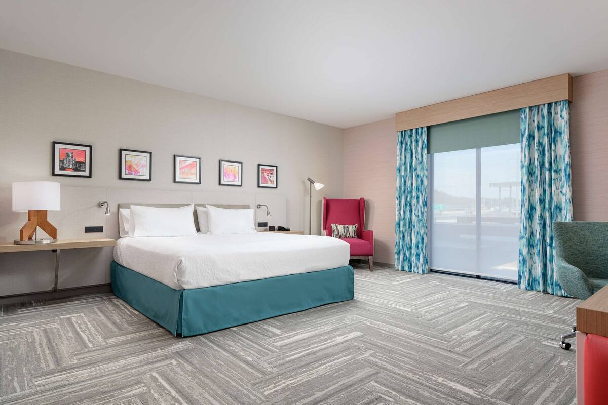 Habitación de hotel con cama, escritorio con lámpara, silla y un ventanal con cortinas.