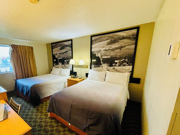 A hotel room with two beds, a desk, a chair, a window with curtains, and a large landscape painting on the wall.