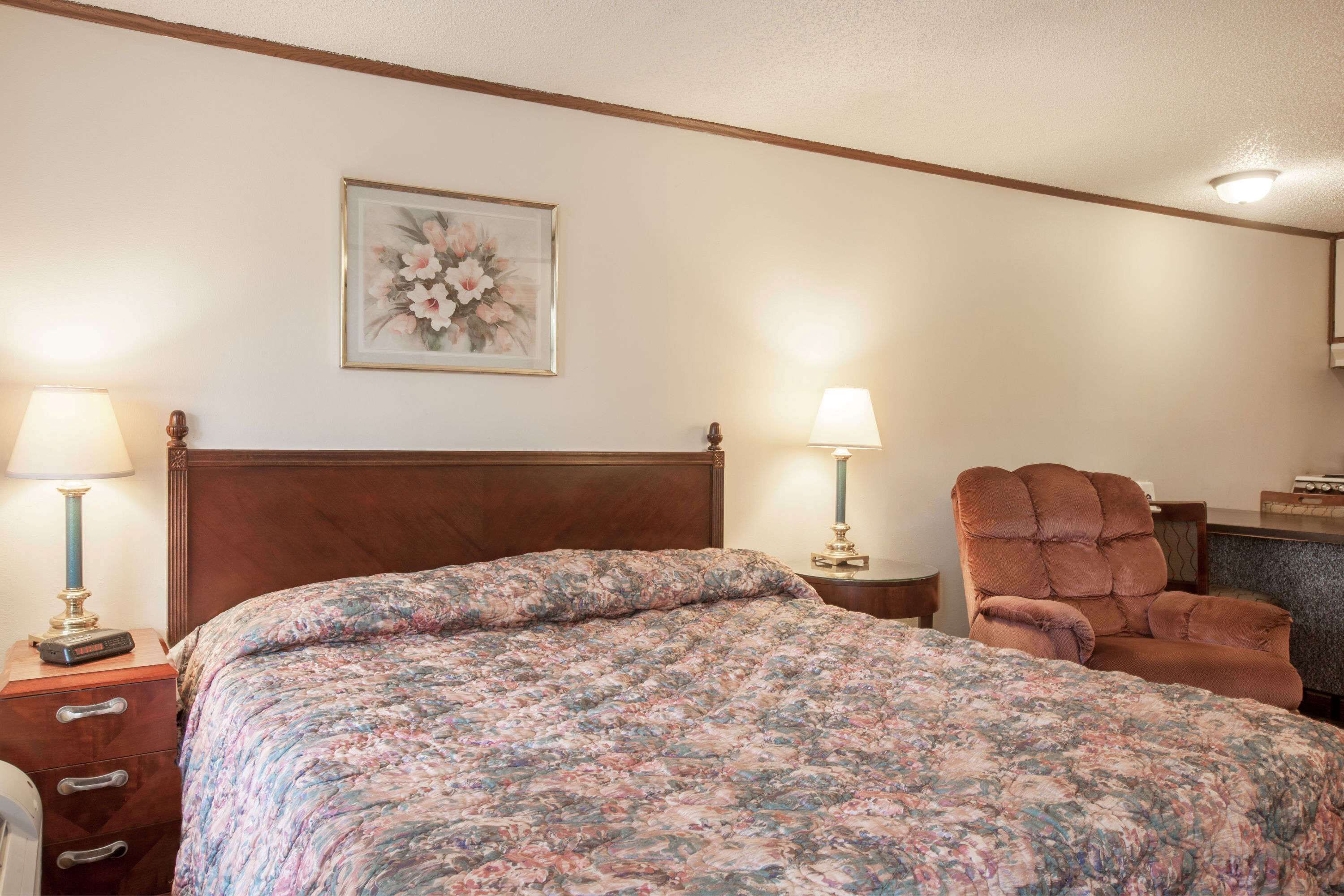 A neatly made bed with a floral bedspread, two bedside lamps, a wooden headboard, and a chair in a hotel room.