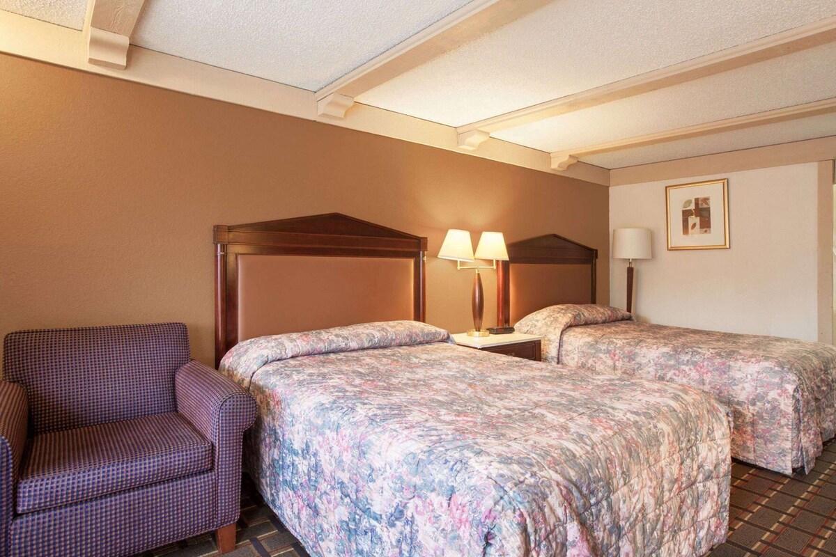 Una habitación de hotel con un sillón a cuadros, dos camas con ropa de cama floral, un cabecero de madera, dos lámparas de noche y un cuadro en la pared.