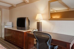 Una habitación de hotel con un escritorio, televisión, lámpara, silla y espejo.